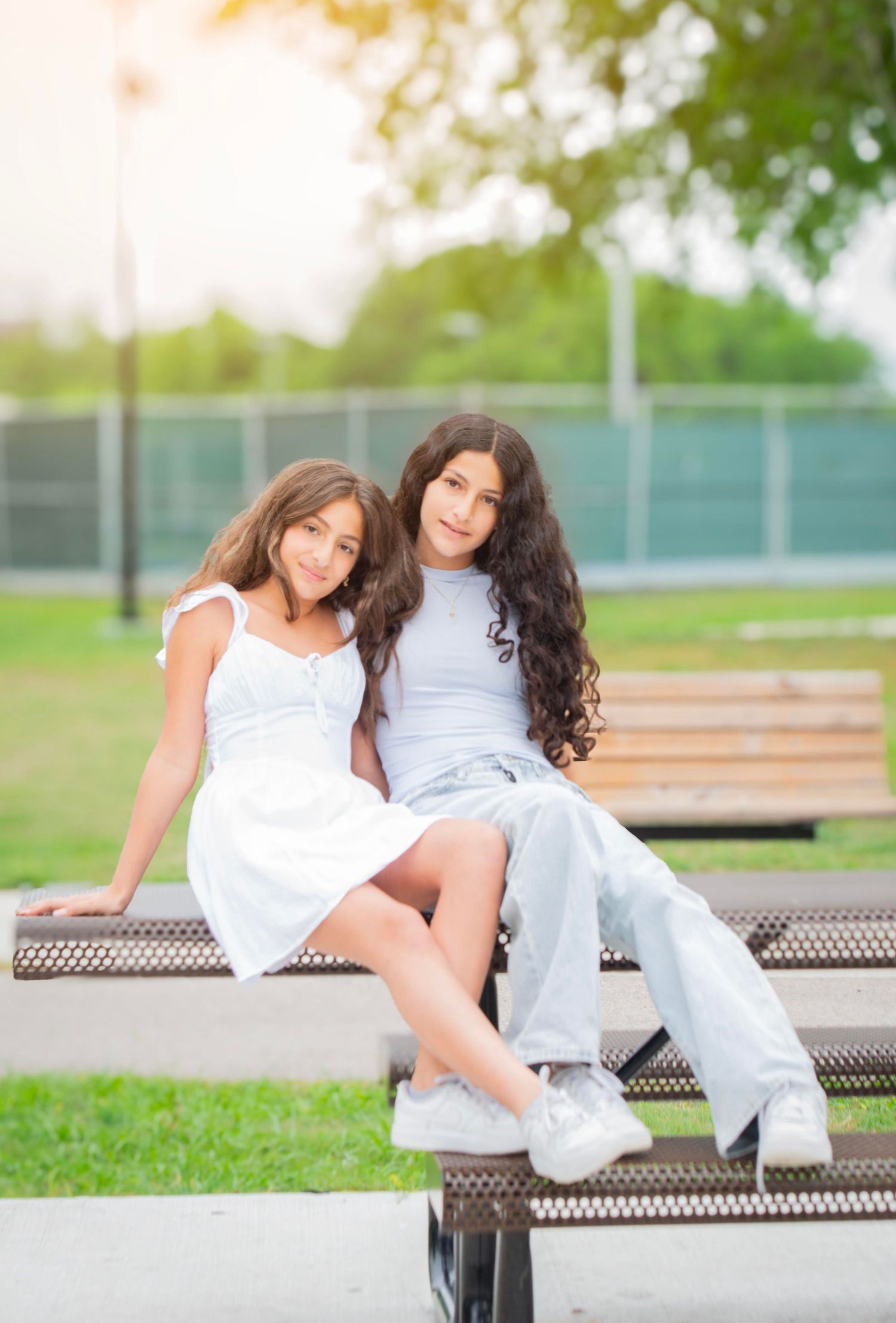 Two young girls in white and light blue outfits sitting together on a park bench in sunlight.