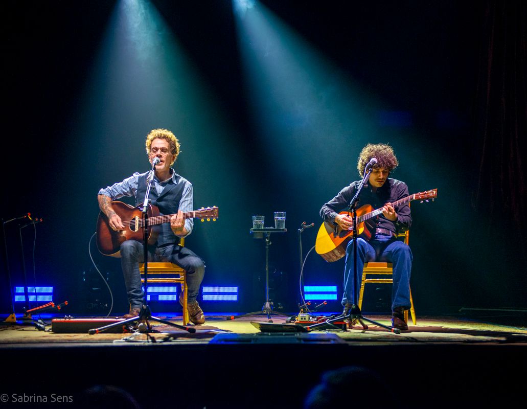 Two acoustic guitarists perform seated on a dimly lit stage with dramatic blue and green spotlights overhead.