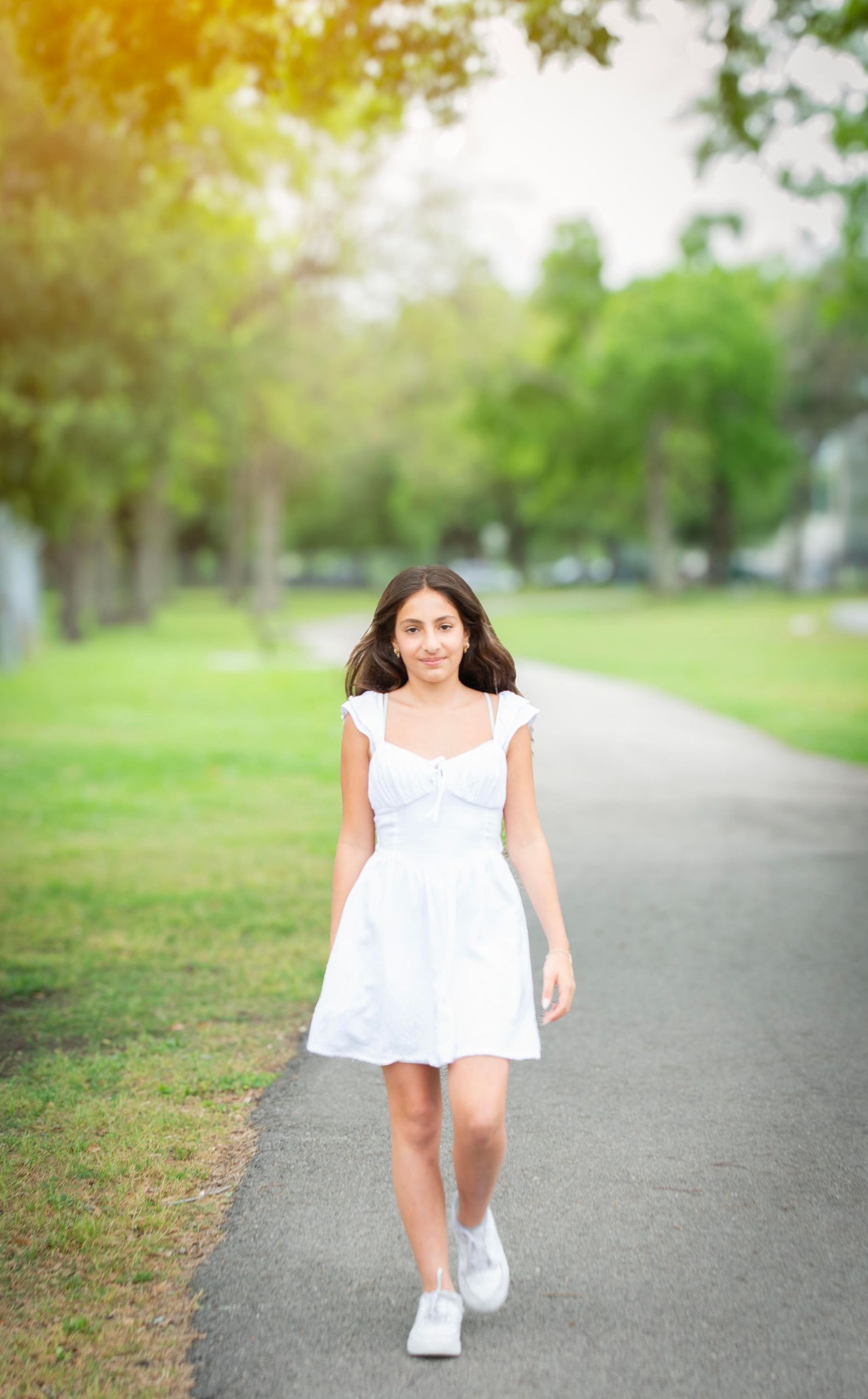Young woman in white dress and sneakers walking on a park pathway surrounded by green trees.