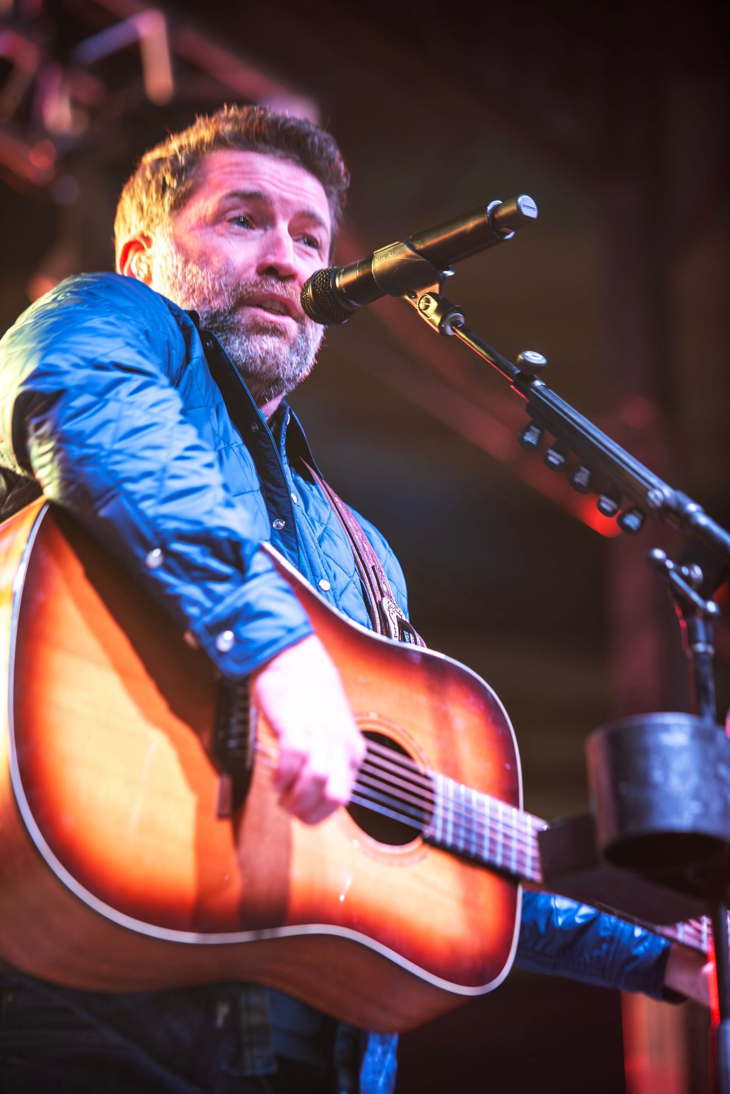 A bearded male guitarist in a blue jacket performs at a microphone with an acoustic guitar on stage.