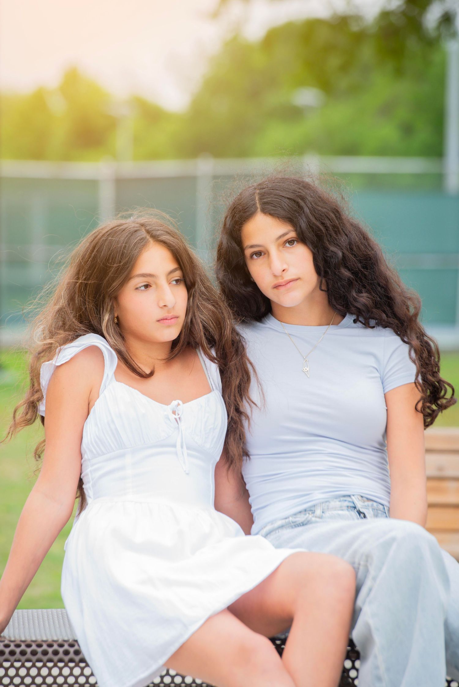 Two young girls with long wavy hair sitting together outdoors, wearing casual summer outfits near a tennis court.