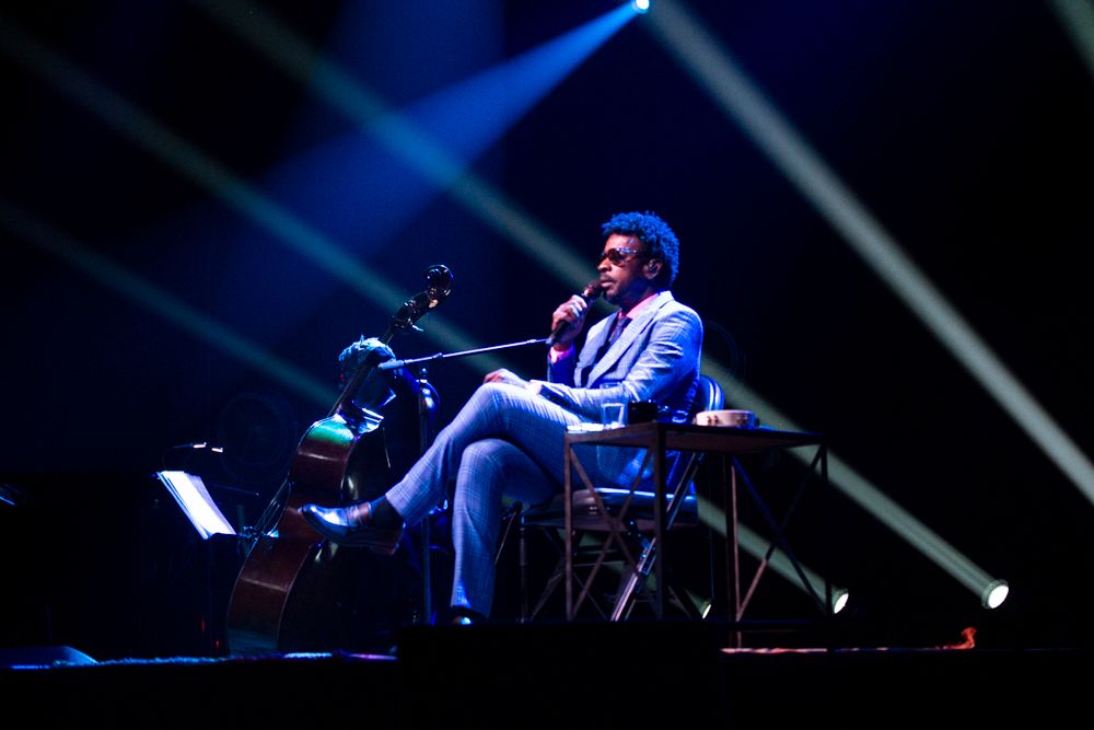 Male musician in blue suit seated playing keyboard on stage with dramatic light beams.