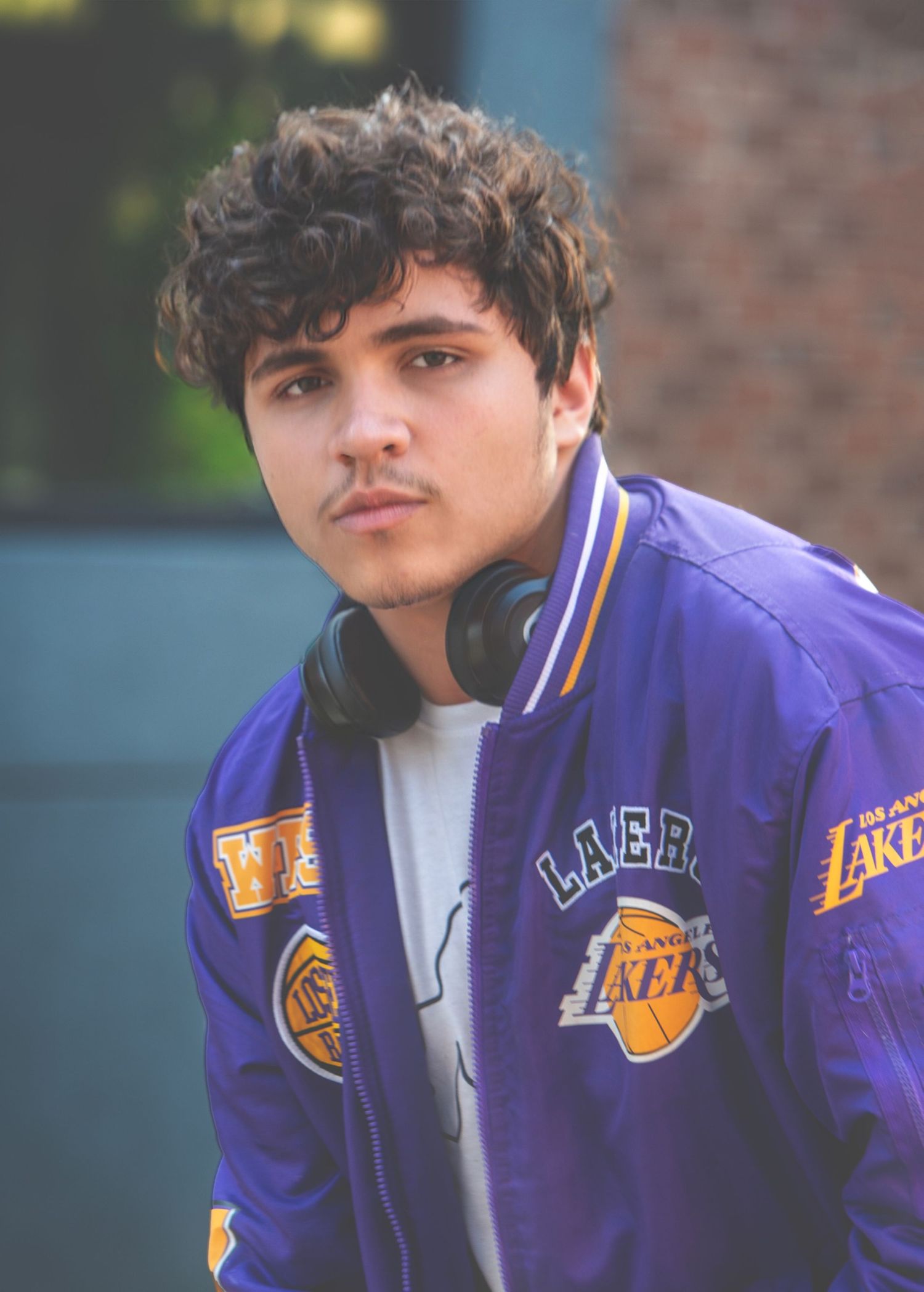 Young man with curly hair wearing purple LA Lakers jacket and headphones around neck in urban setting.