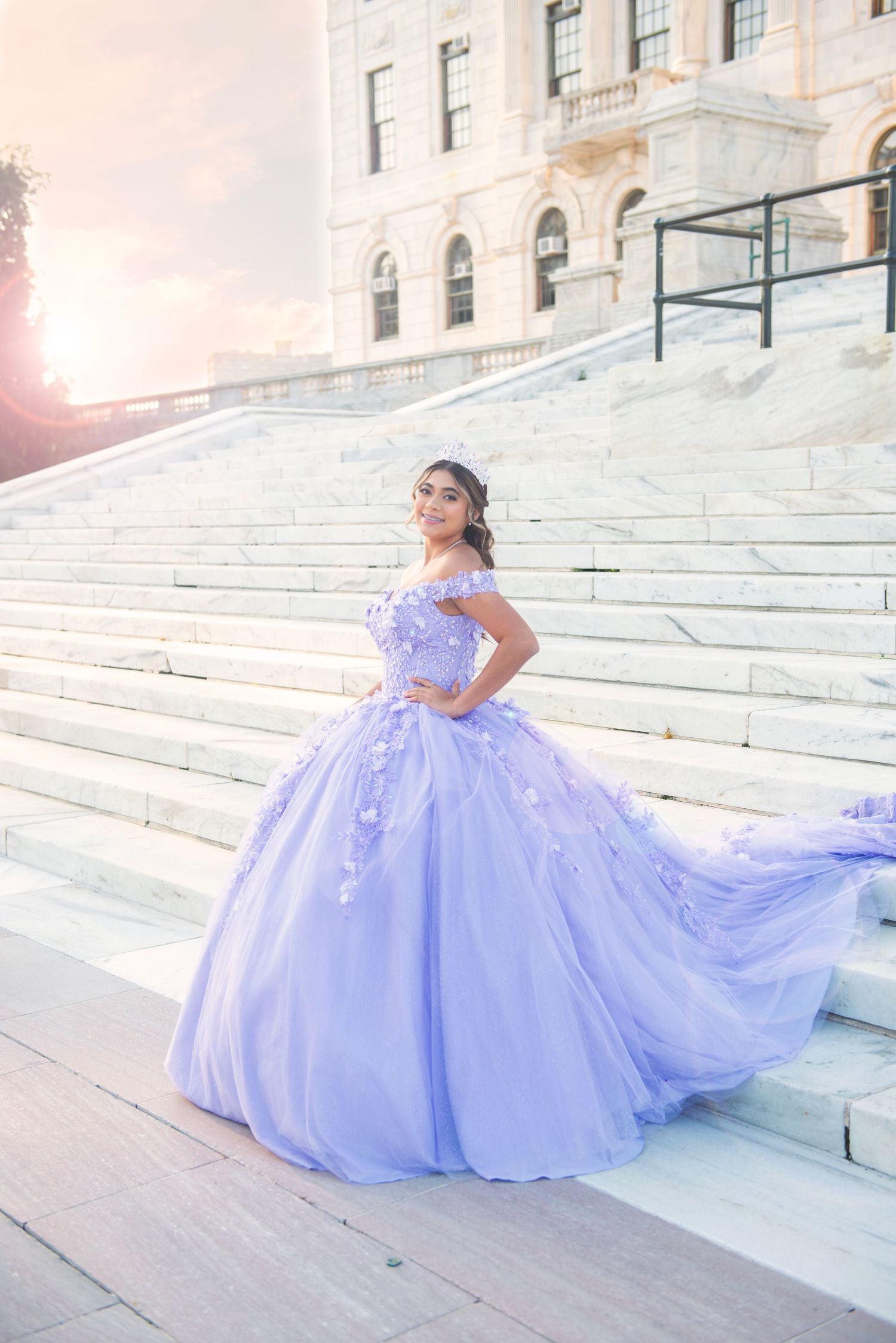 Young woman in a flowing lavender quinceañera ball gown posing on grand marble steps at sunset.