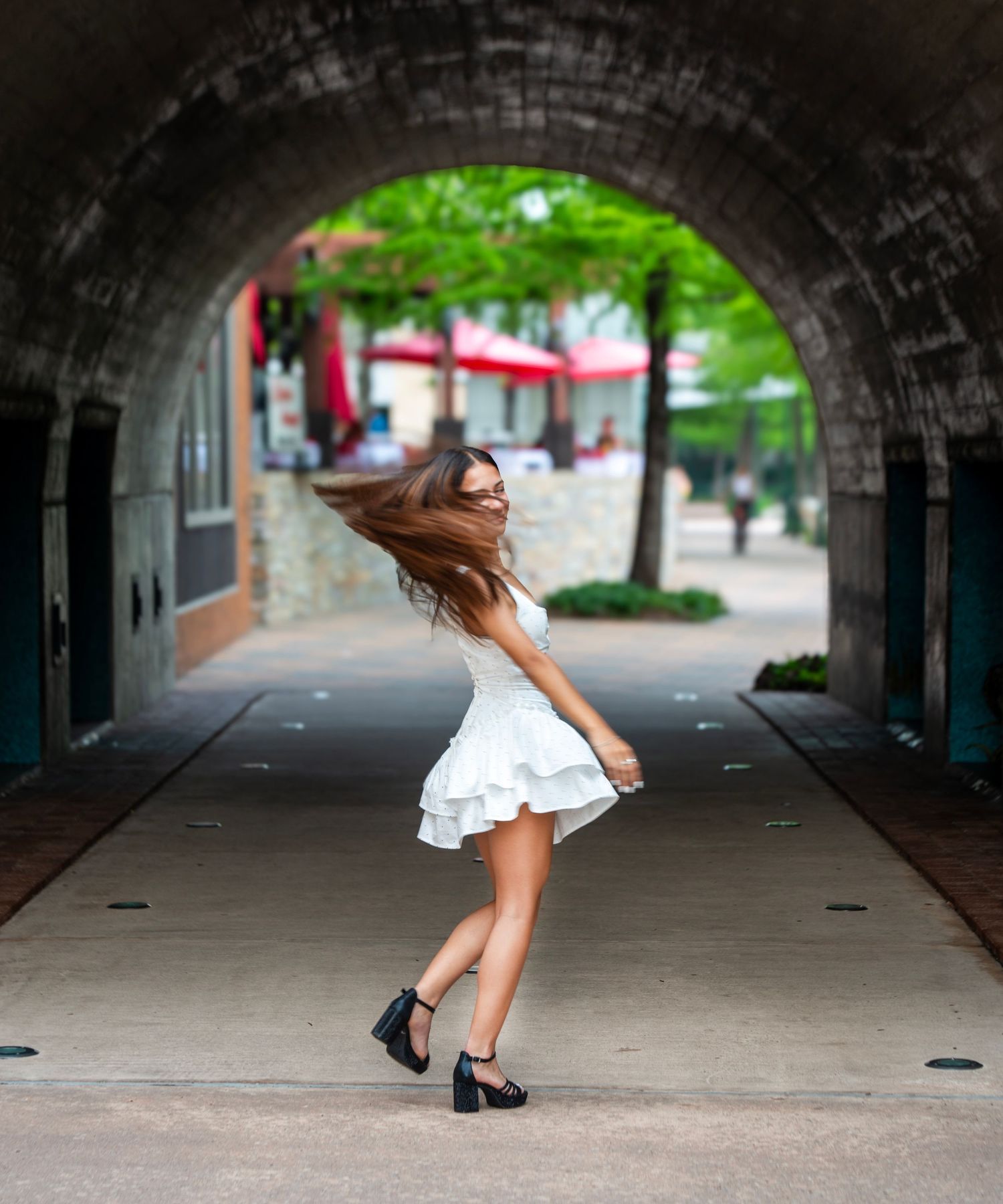 Young woman in white ruffled dress twirling under stone arch tunnel, hair flowing, wearing black heels outdoors.