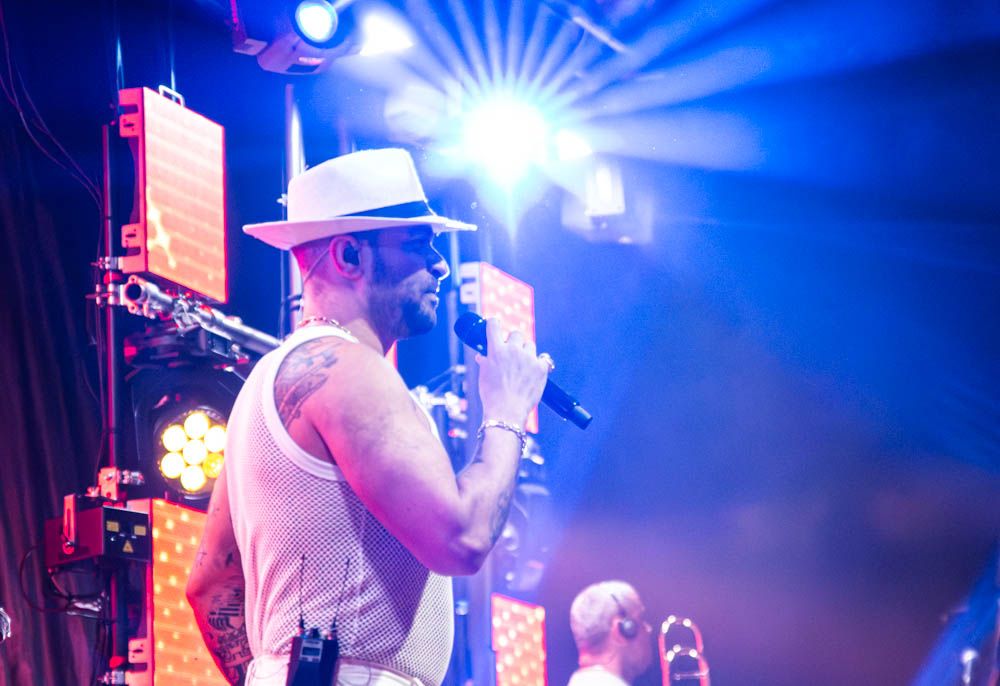 A male vocalist in a white fedora hat sings passionately into a microphone under blue stage lighting.