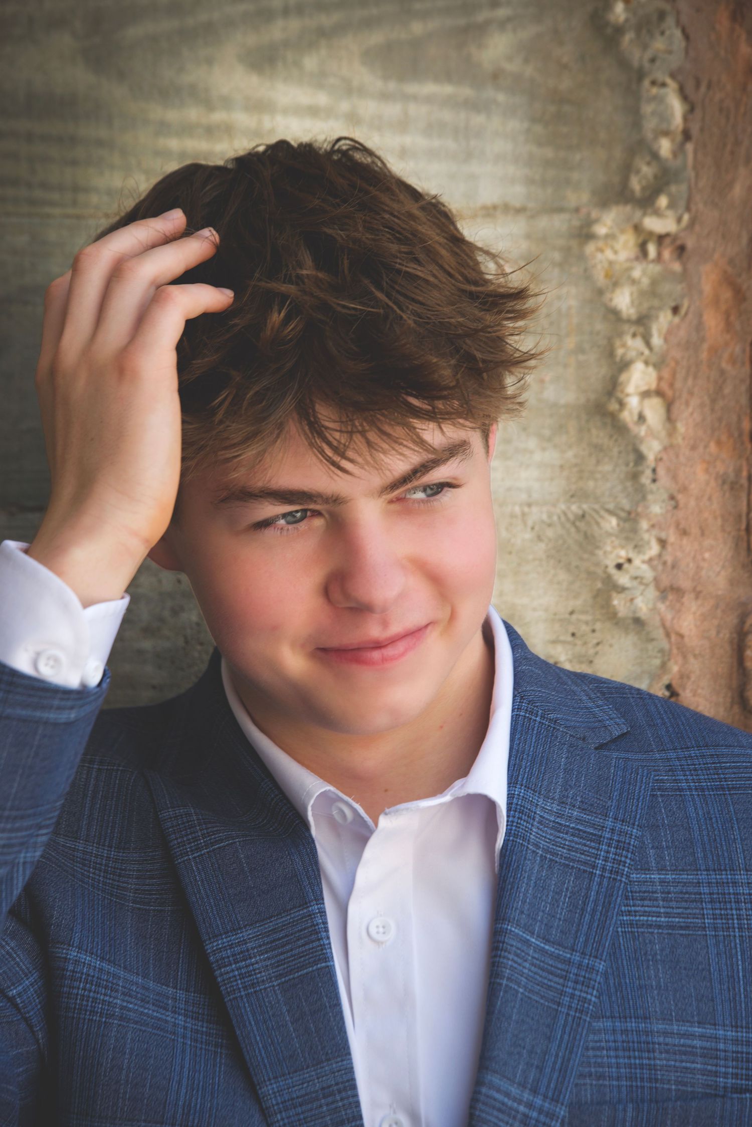 Young man in blue plaid blazer smiling, hand in brown hair, against rustic wall background.