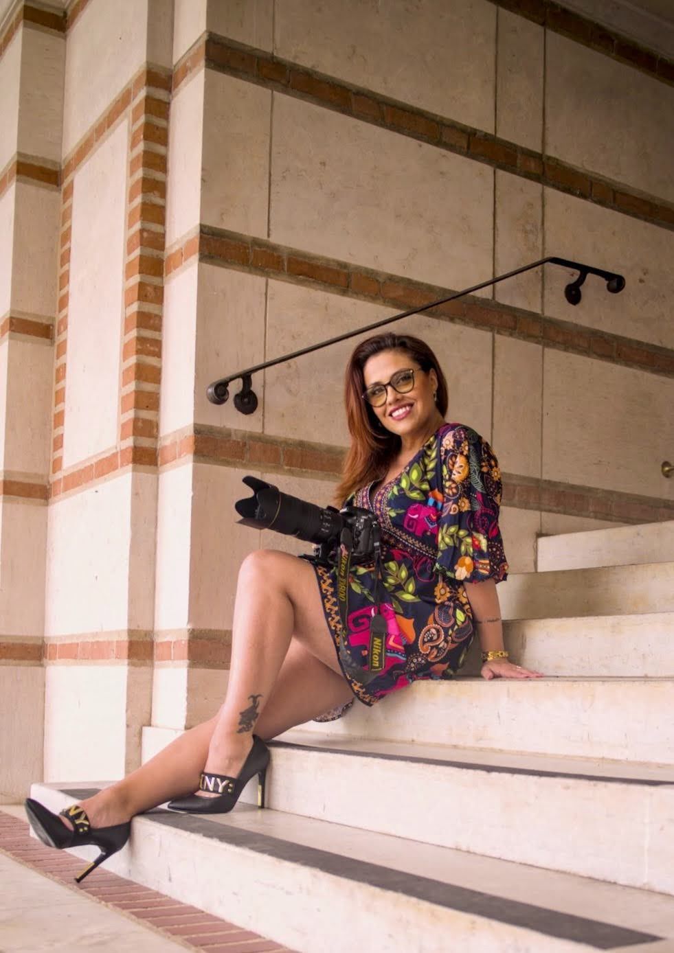 Stylish woman in floral dress and heels sitting on marble steps holding a camera, wearing glasses and smiling.