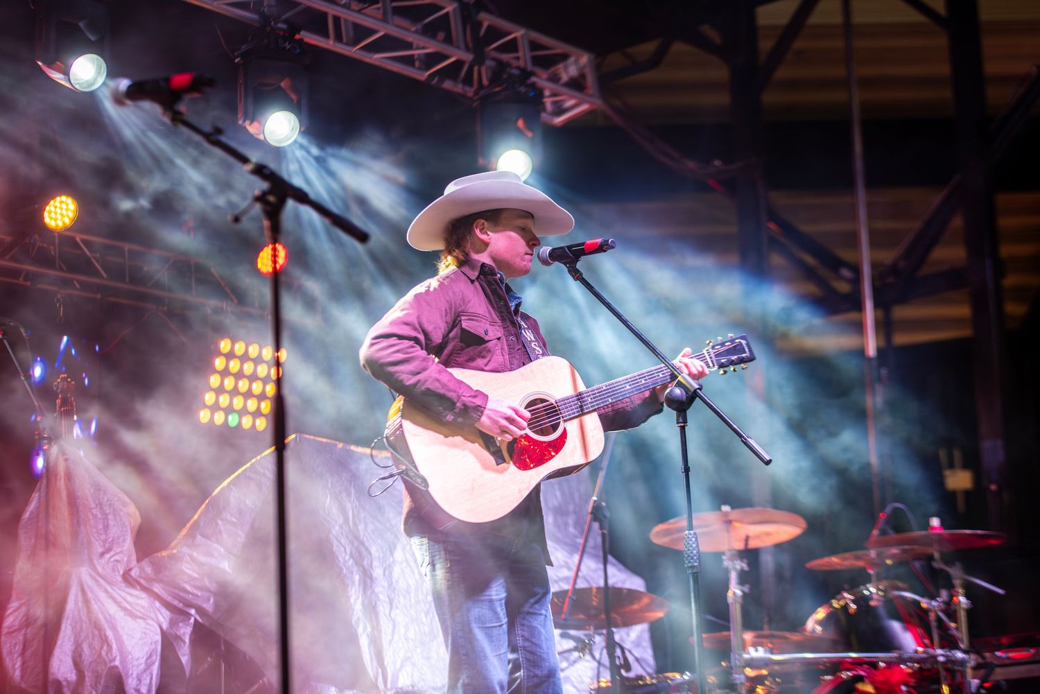 Country singer performing on smoky stage with acoustic guitar wearing white cowboy hat and jeans.