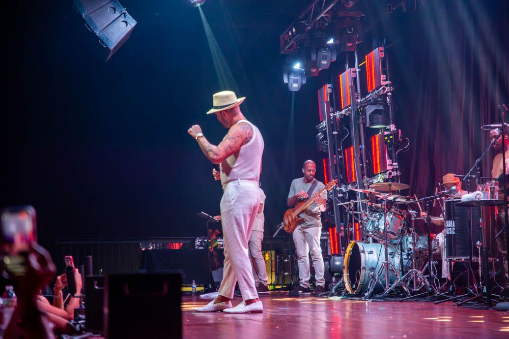 A male singer in white hat and outfit performs energetically on stage with a full band and dramatic lighting.