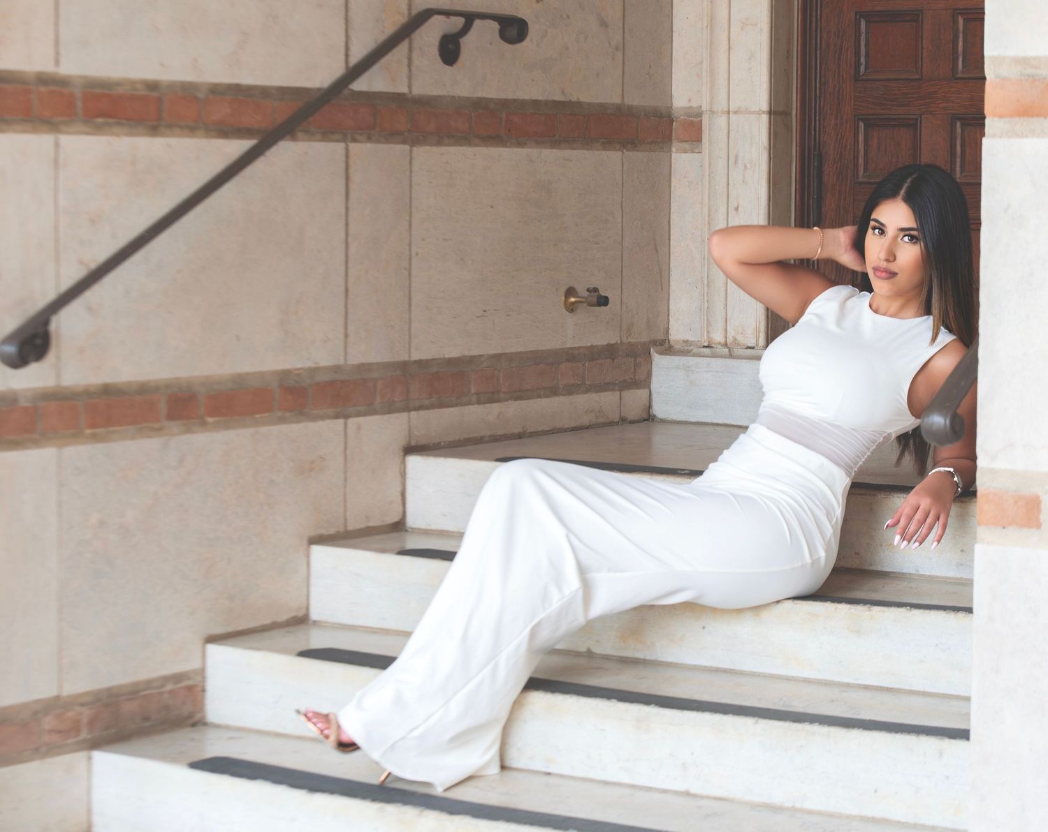 Woman in white outfit sitting gracefully on marble steps, posing with hand behind head in an elegant architectural setting.