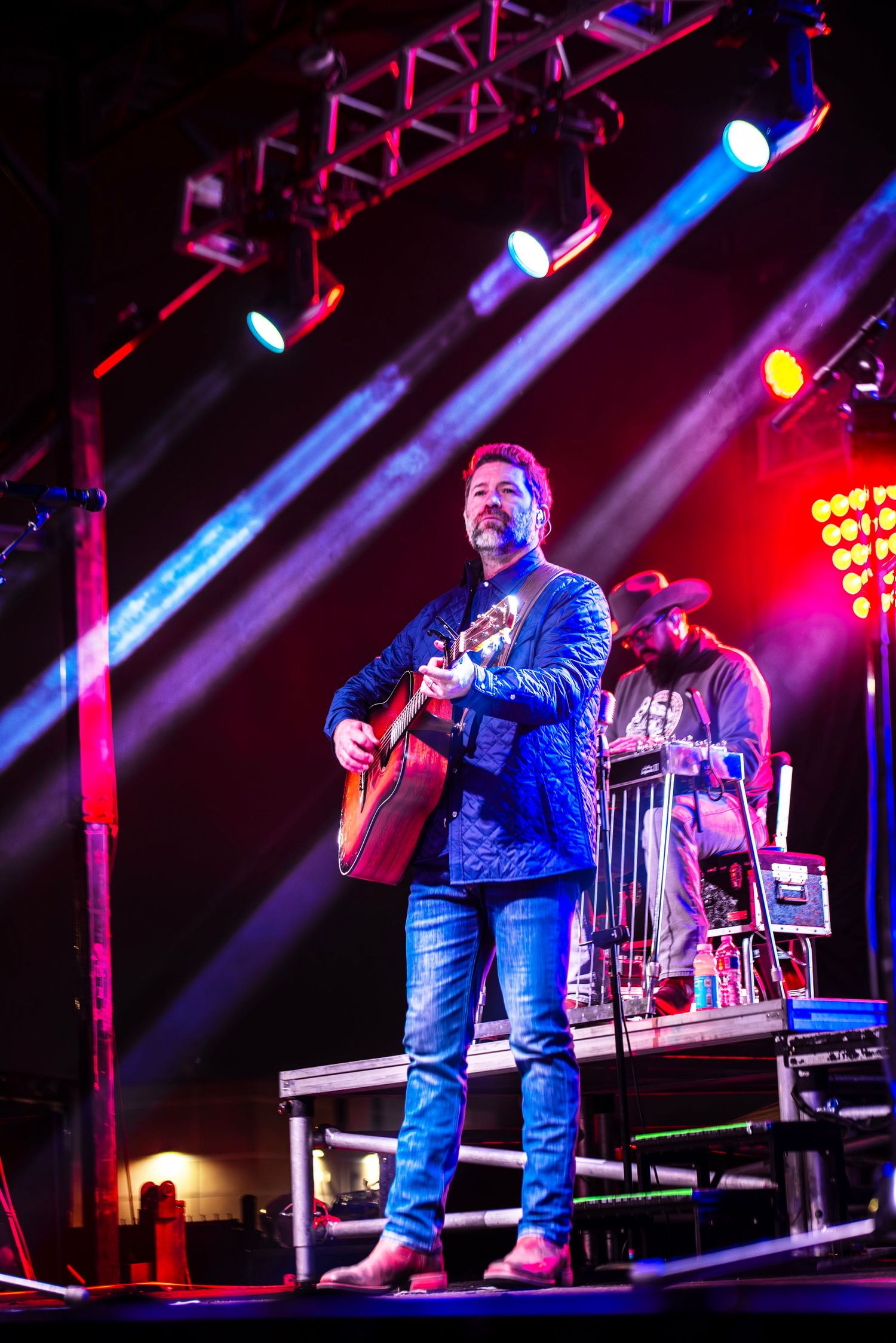 Male guitarist in blue blazer performing on colorful illuminated concert stage with band.