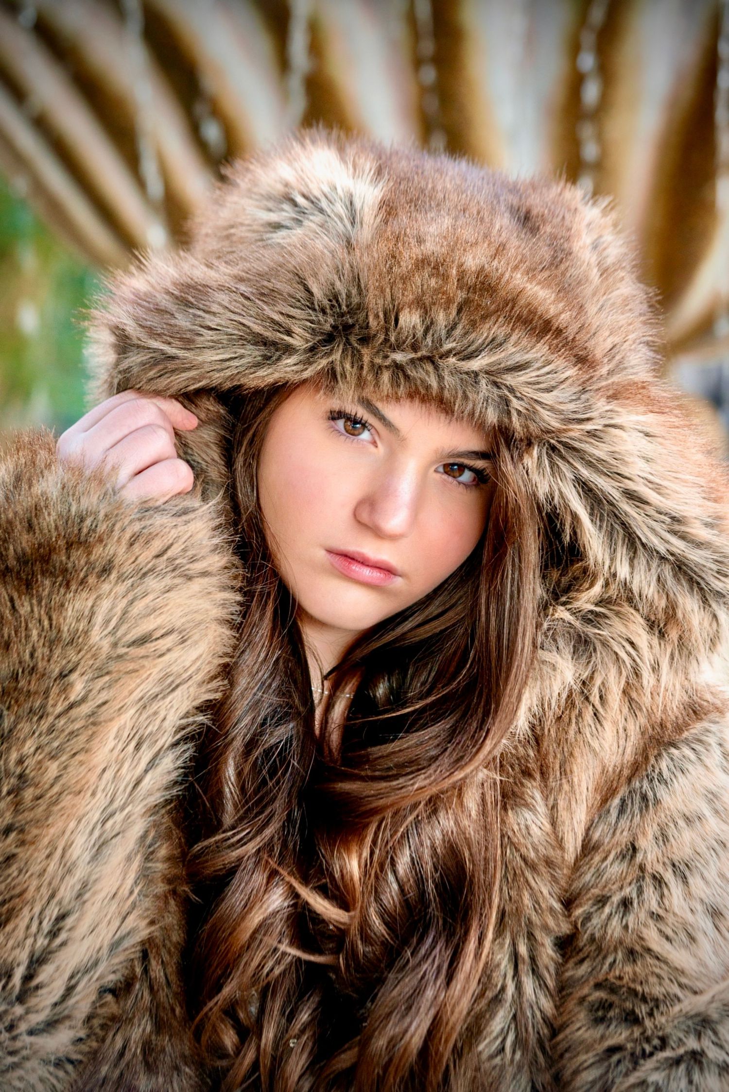 Young woman with long brown hair wearing a hooded faux fur coat posing with a serious expression.