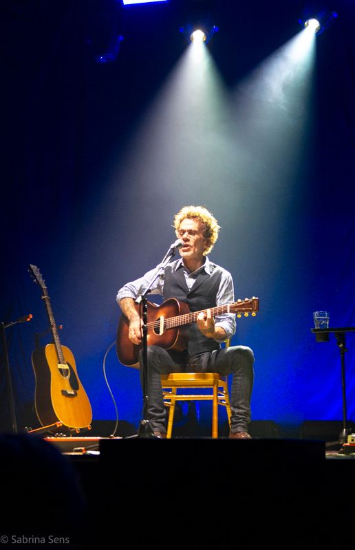 Curly-haired musician seated on stool playing acoustic guitar on stage under blue spotlight.