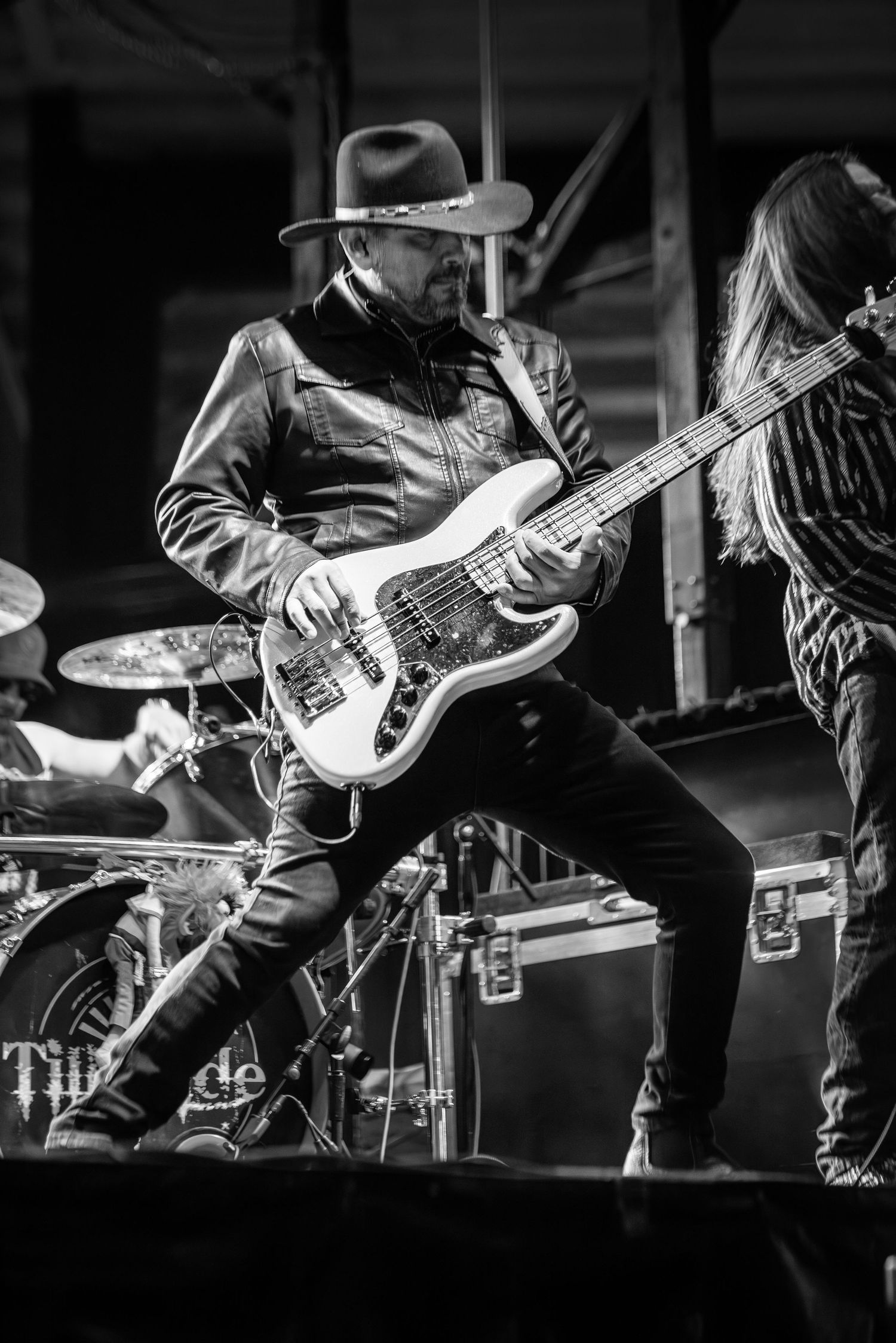 Black and white image of a bassist in a cowboy hat playing a white bass guitar on stage at a live show.