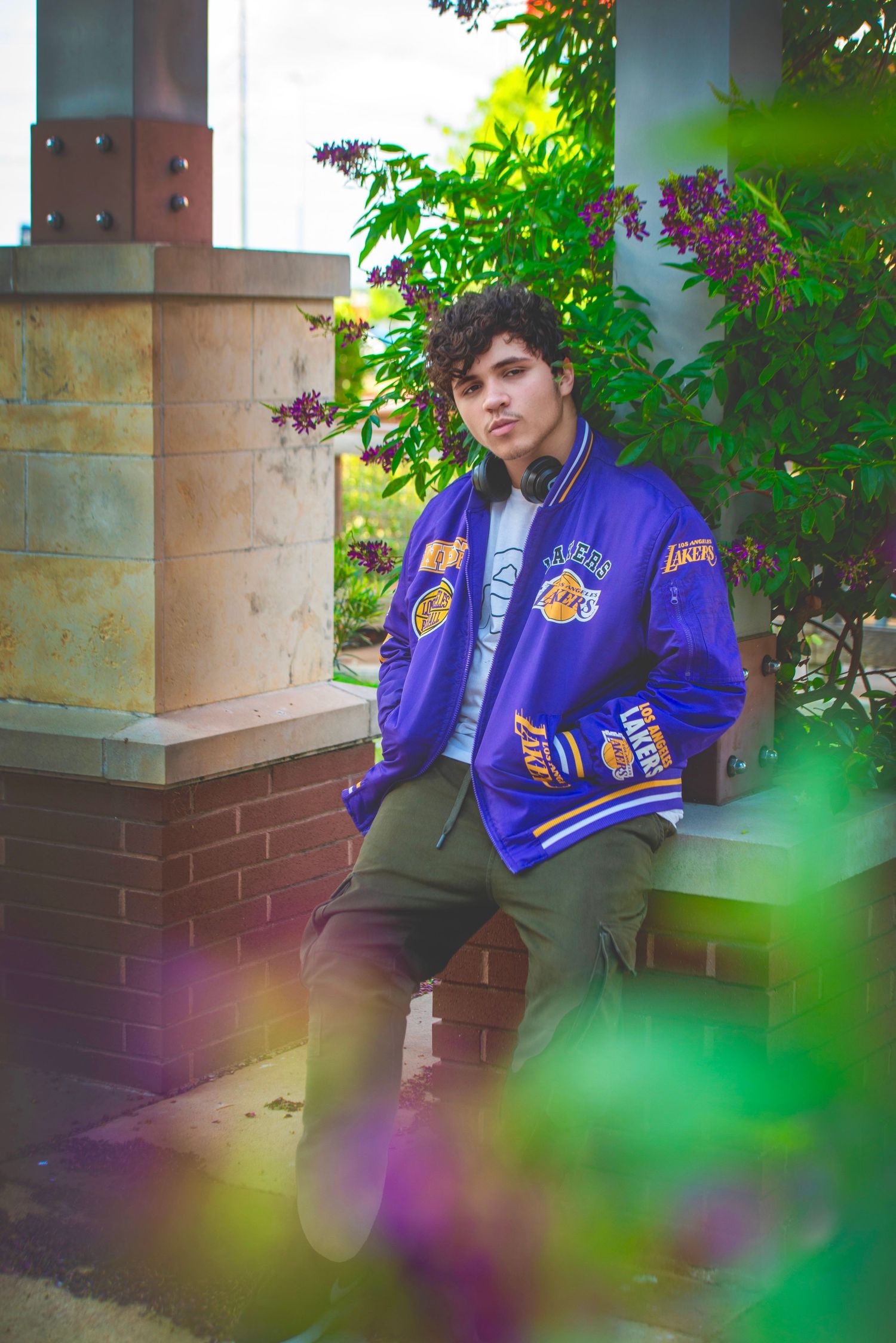 Teen boy in purple LA Lakers varsity jacket sitting on steps surrounded by lush green and purple garden foliage.
