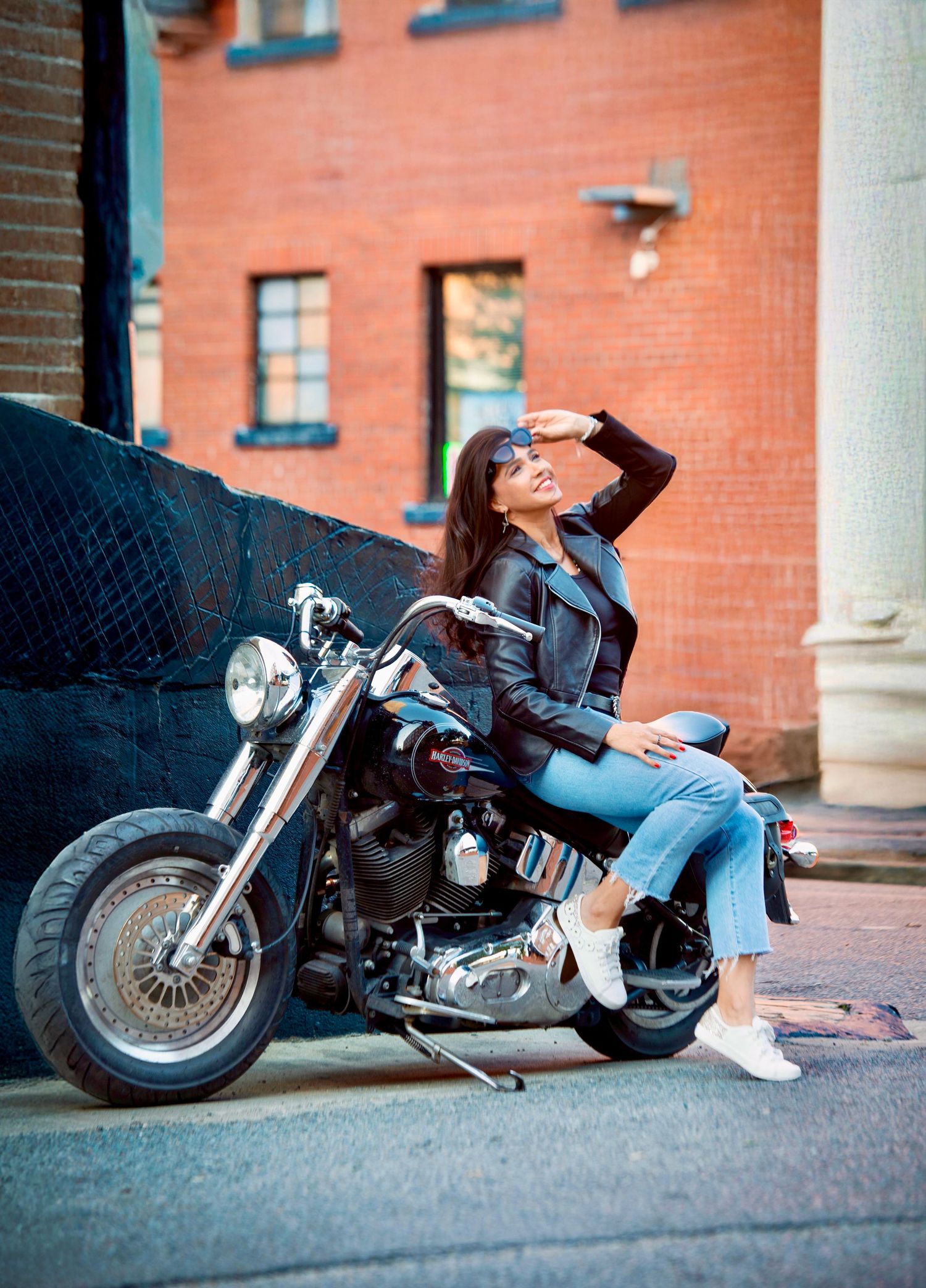 Woman in leather jacket and jeans posing on a Harley-Davidson motorcycle on an urban street.