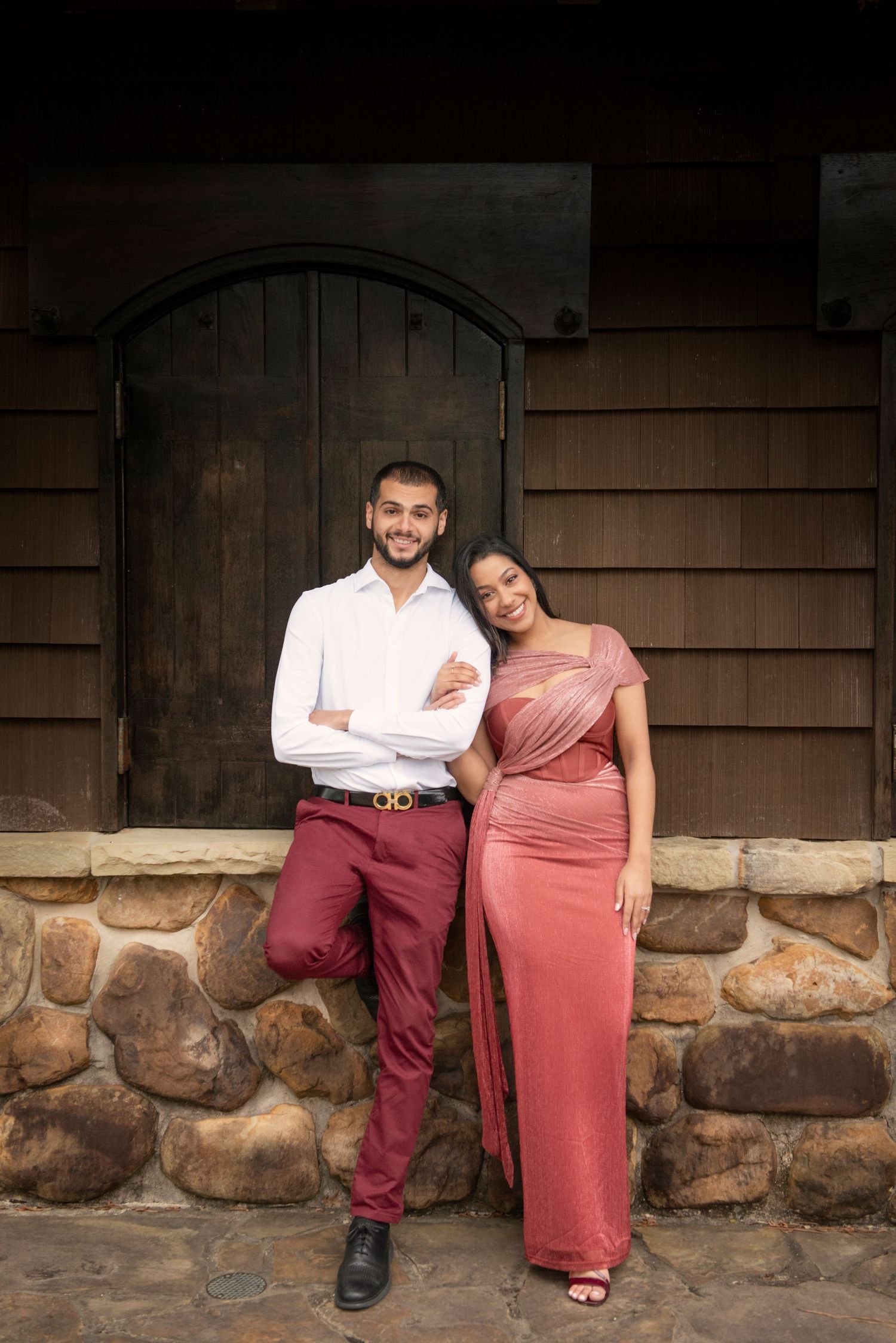 Elegant couple in coordinating rose and burgundy outfits posing together in front of a wooden arched door.