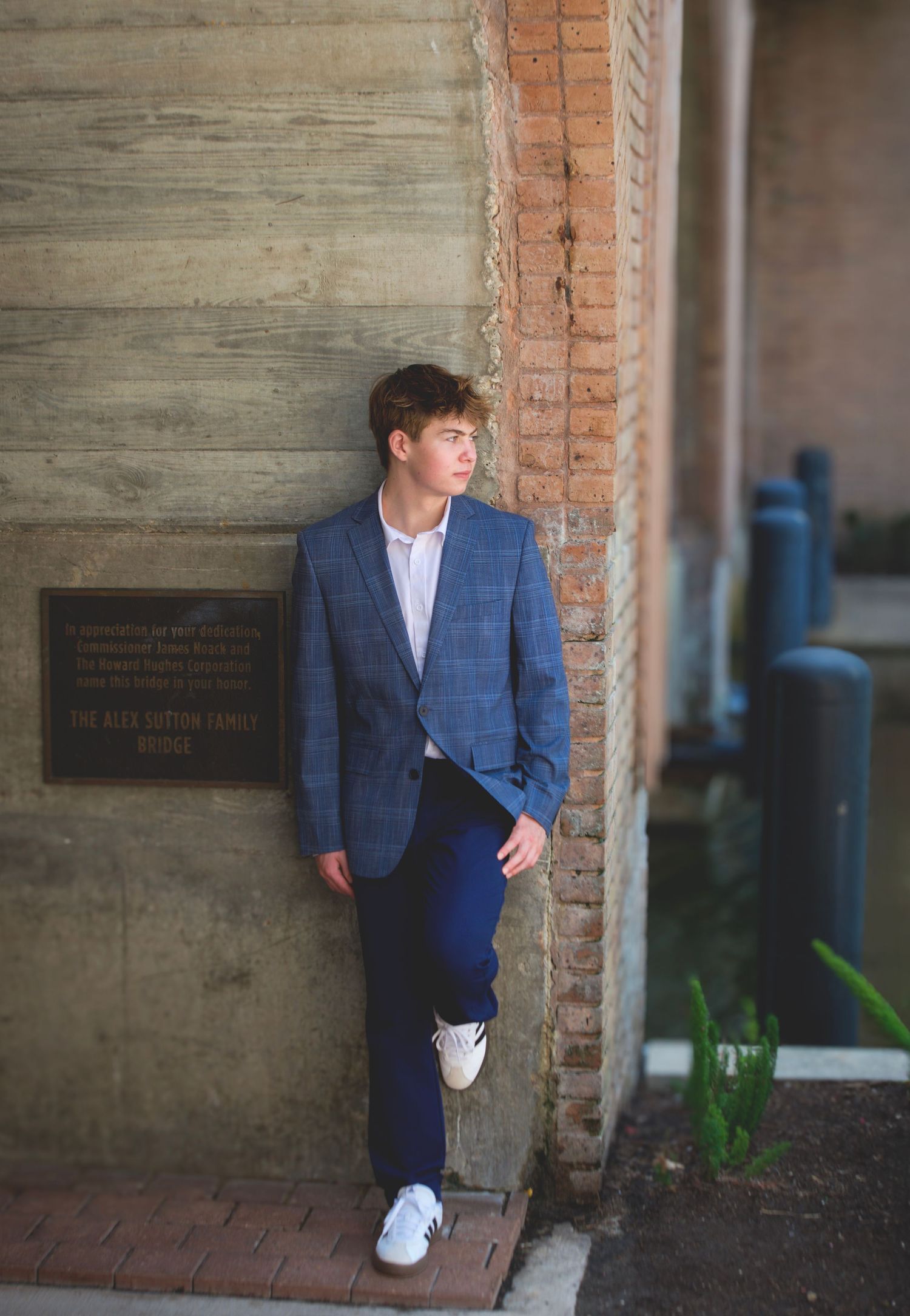 Young man in blue plaid blazer and navy pants leaning against brick wall outdoors.