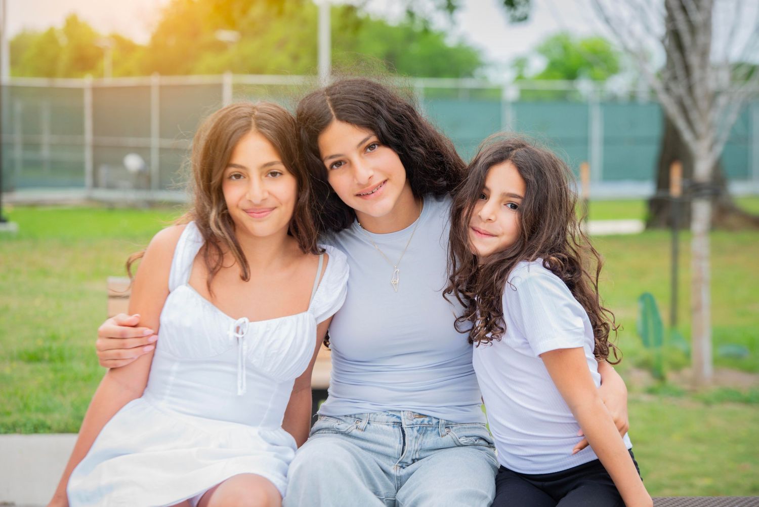Three young girls smiling and hugging outdoors in a sunny park, wearing casual white and blue outfits.