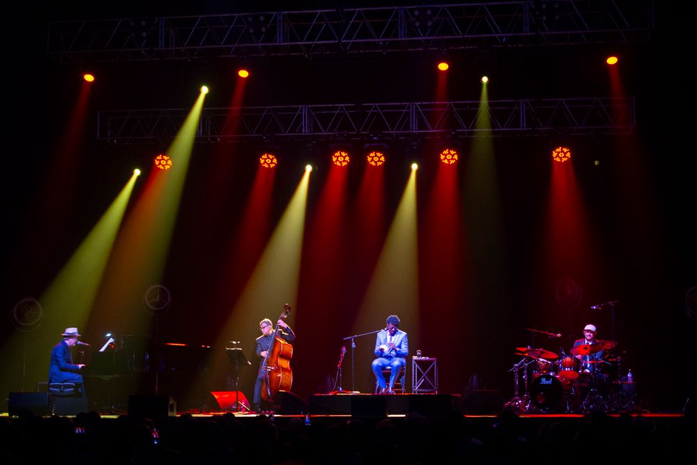 Four-piece band performing on stage under red and yellow concert lighting.