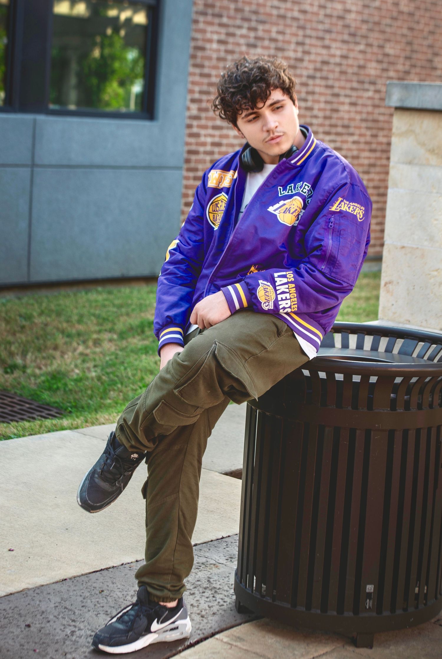 Young man in purple LA Lakers varsity jacket leaning on outdoor trash can in urban environment.