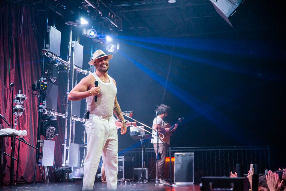 A charismatic male performer in white outfit and hat sings on a colorful lit stage with band members behind him.
