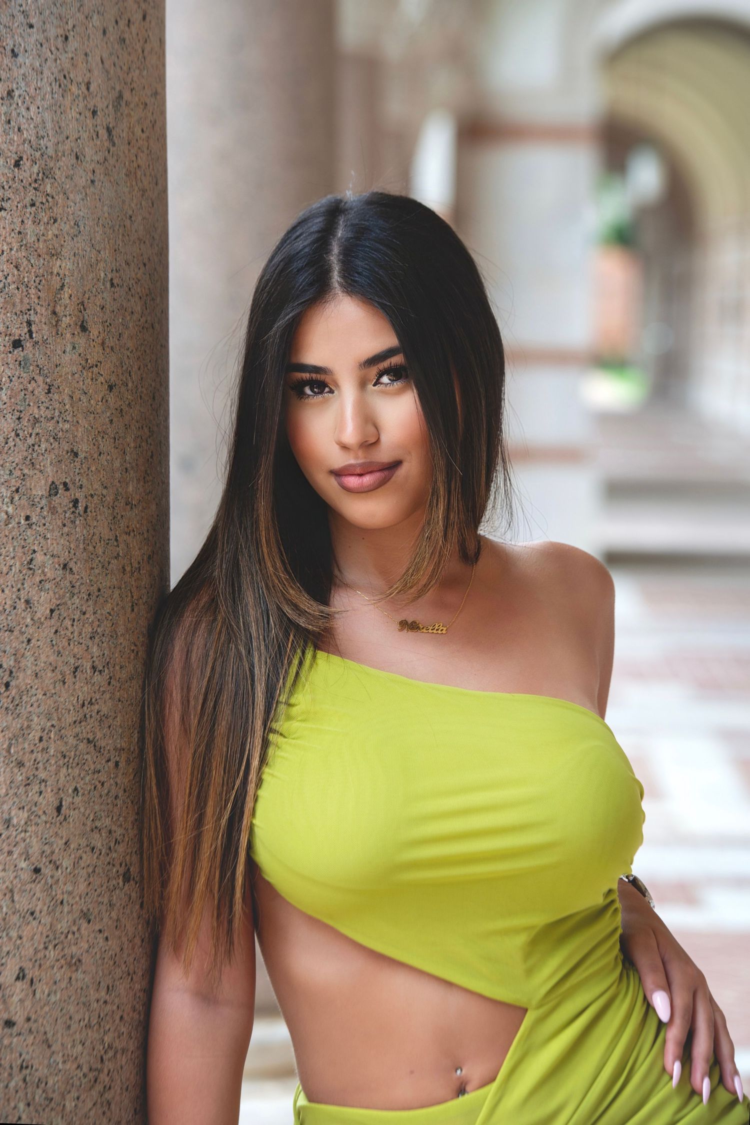 Young woman in yellow-green one-shoulder crop top leaning against stone column in archway.