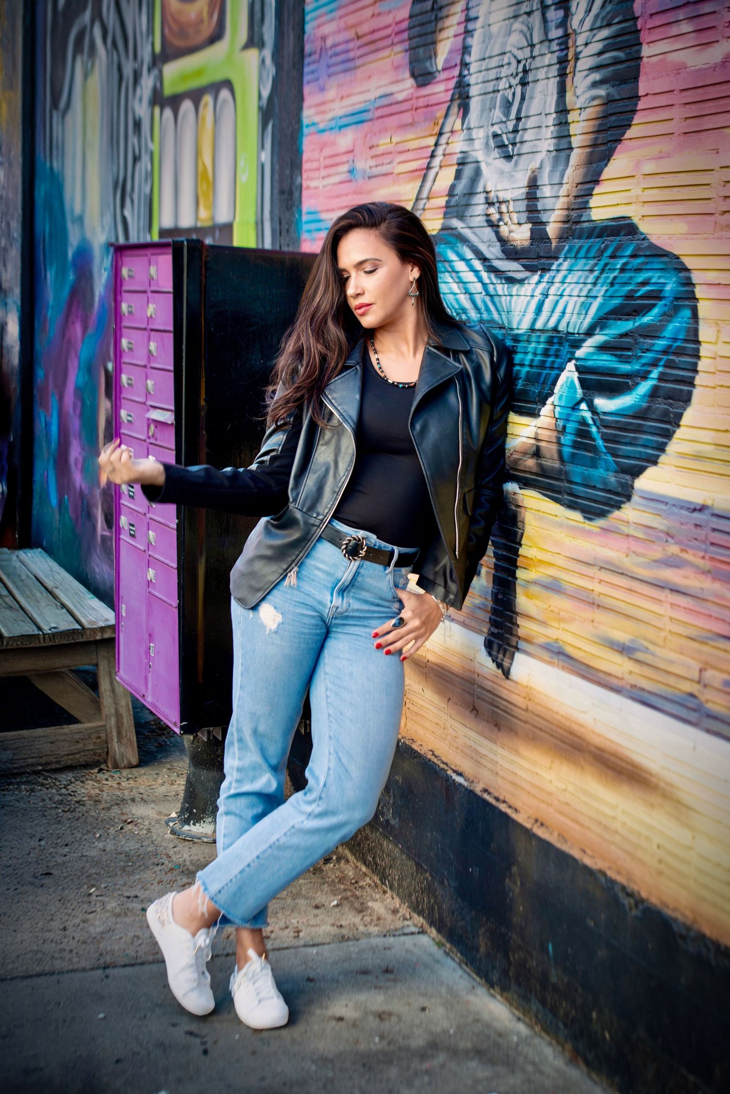 Woman in leather jacket and jeans leaning against a colorful graffiti wall outdoors.