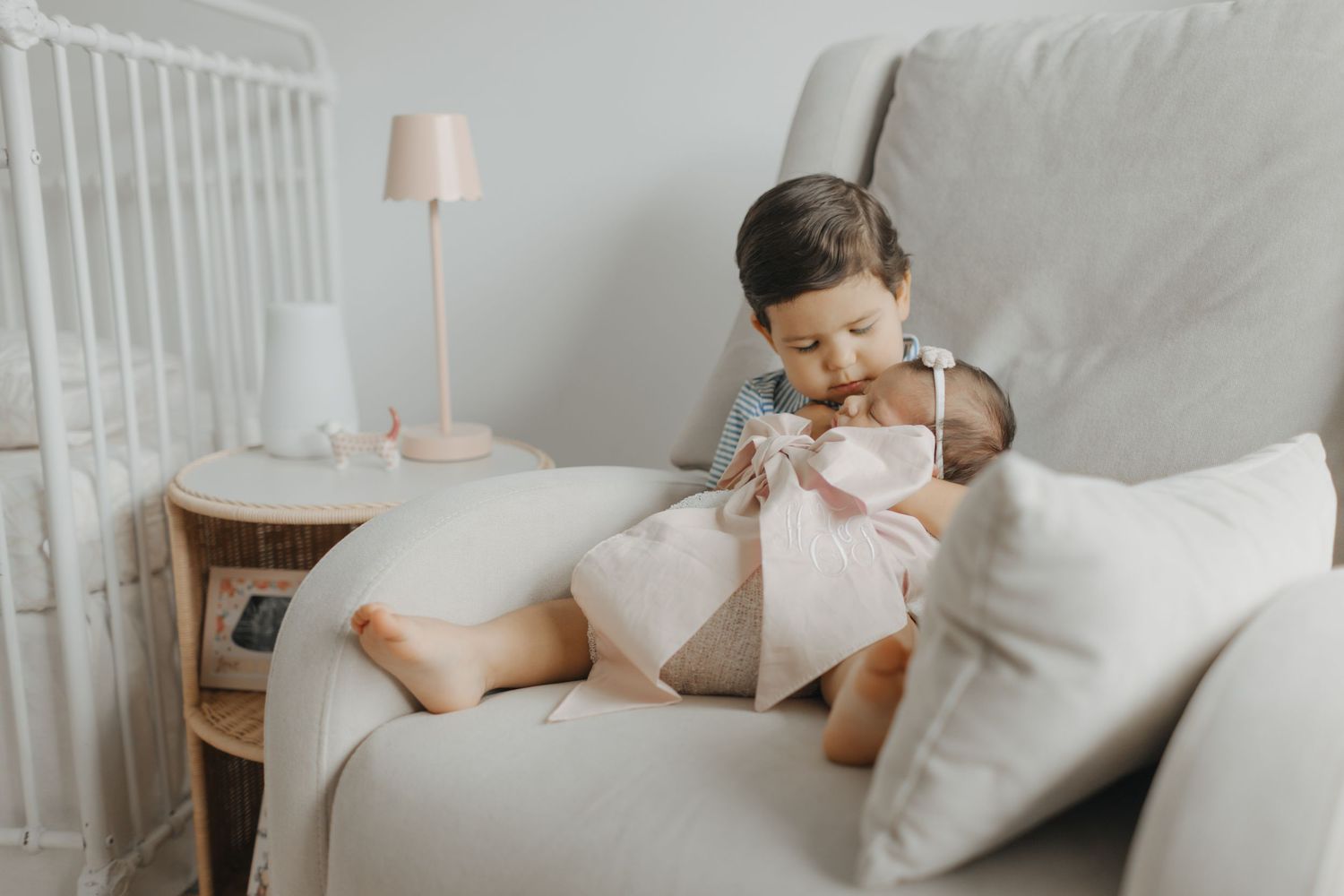 Young toddler boy gently holding a newborn baby in a cozy nursery chair beside a crib and pink lamp.
