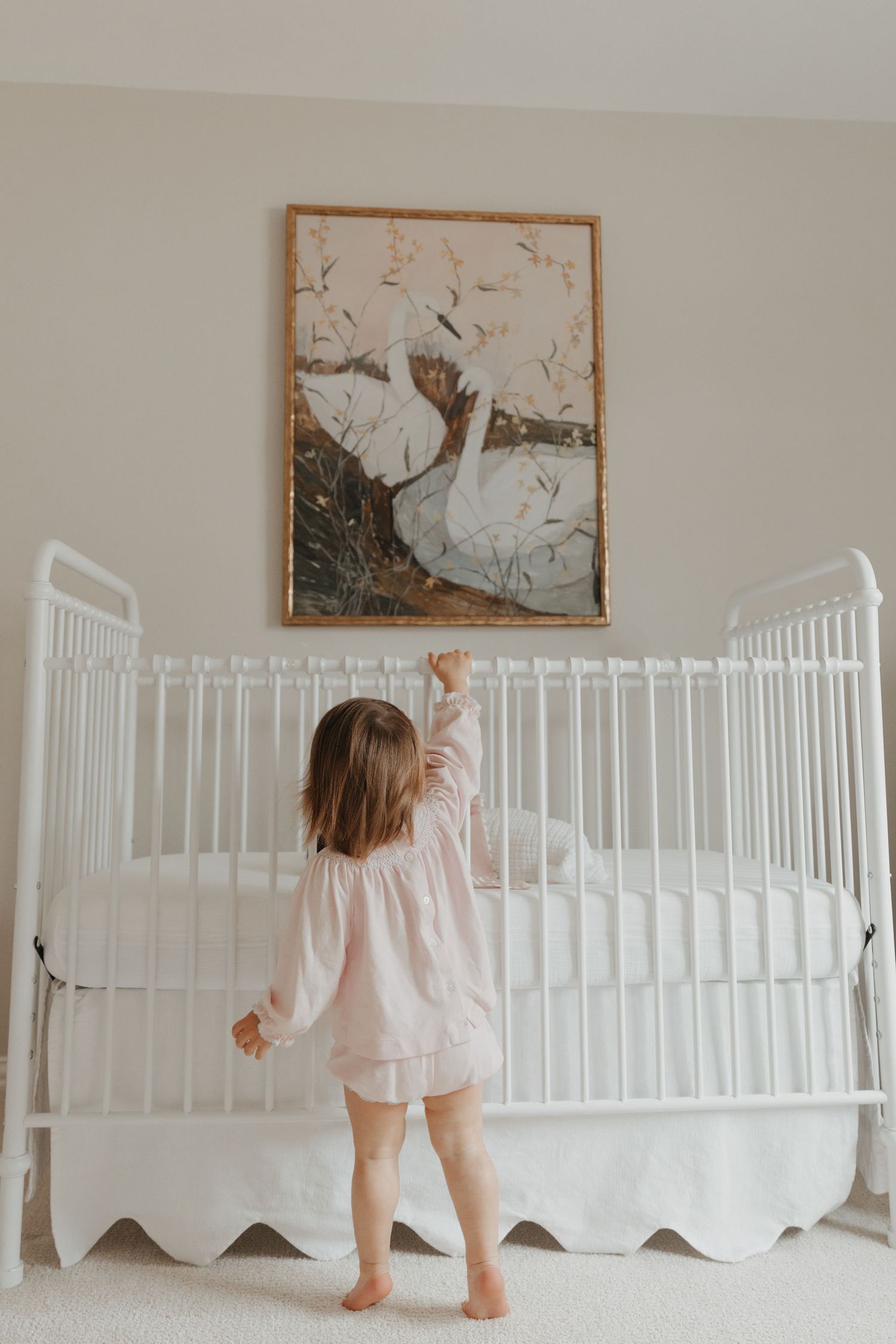 Toddler girl in pink dress standing at a white crib reaching toward a swan painting in a nursery.