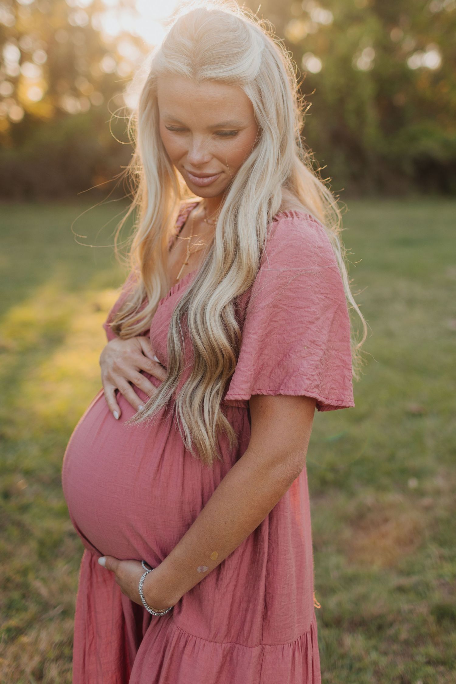 Radiant pregnant woman in rose dress tenderly holding baby bump in warm golden sunlight.