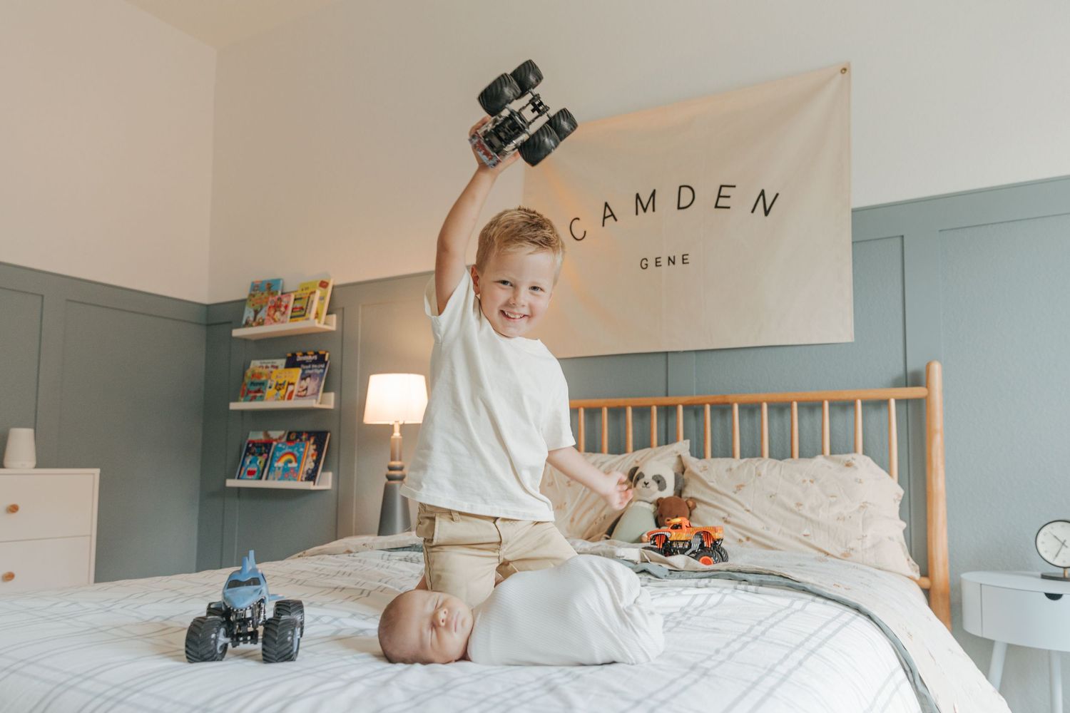 Young boy sits on bed holding toy truck above newborn sibling in room with Camden Gene banner.