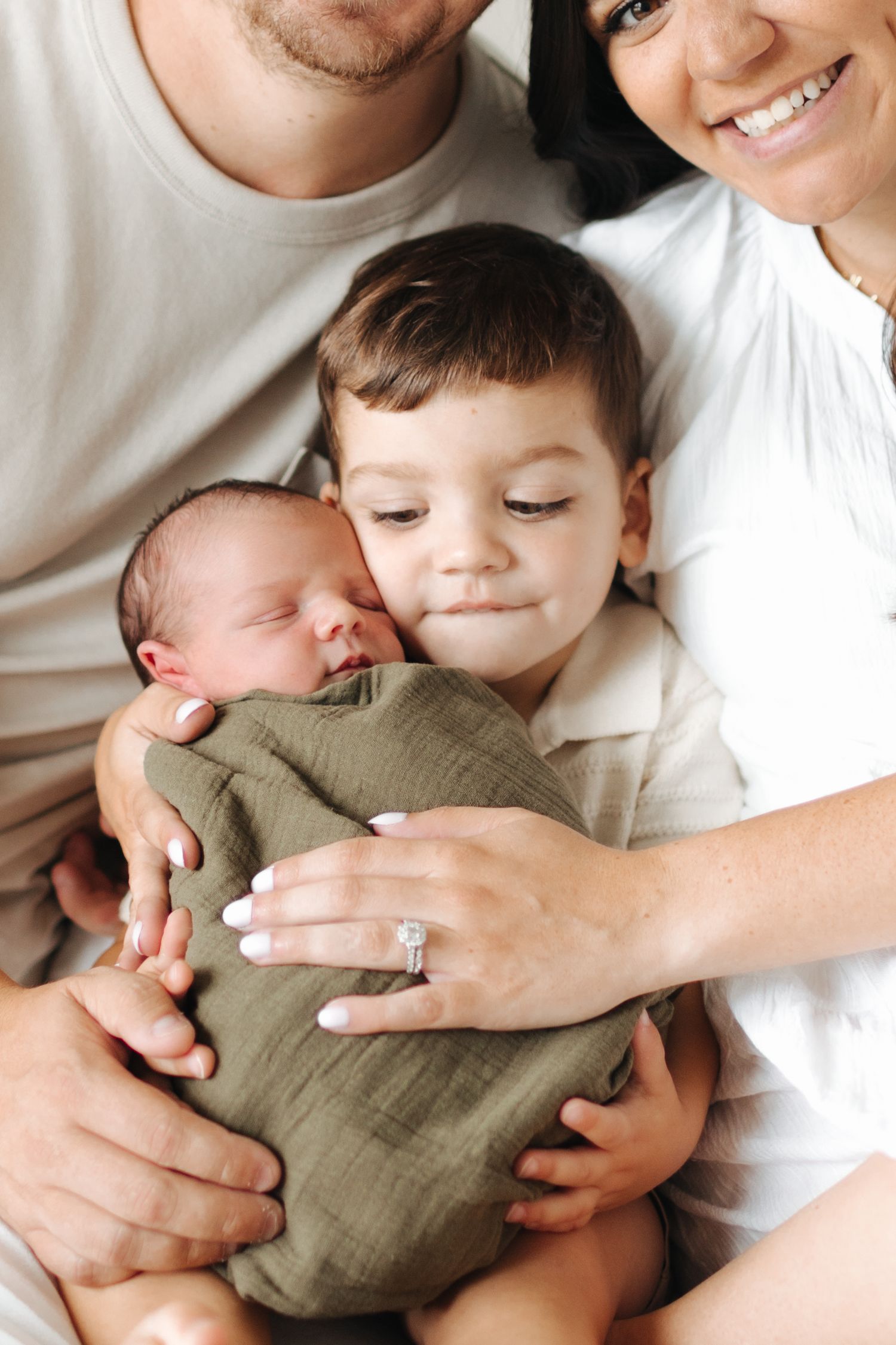 Family portrait with parents and toddler son tenderly holding swaddled newborn baby in olive green wrap.