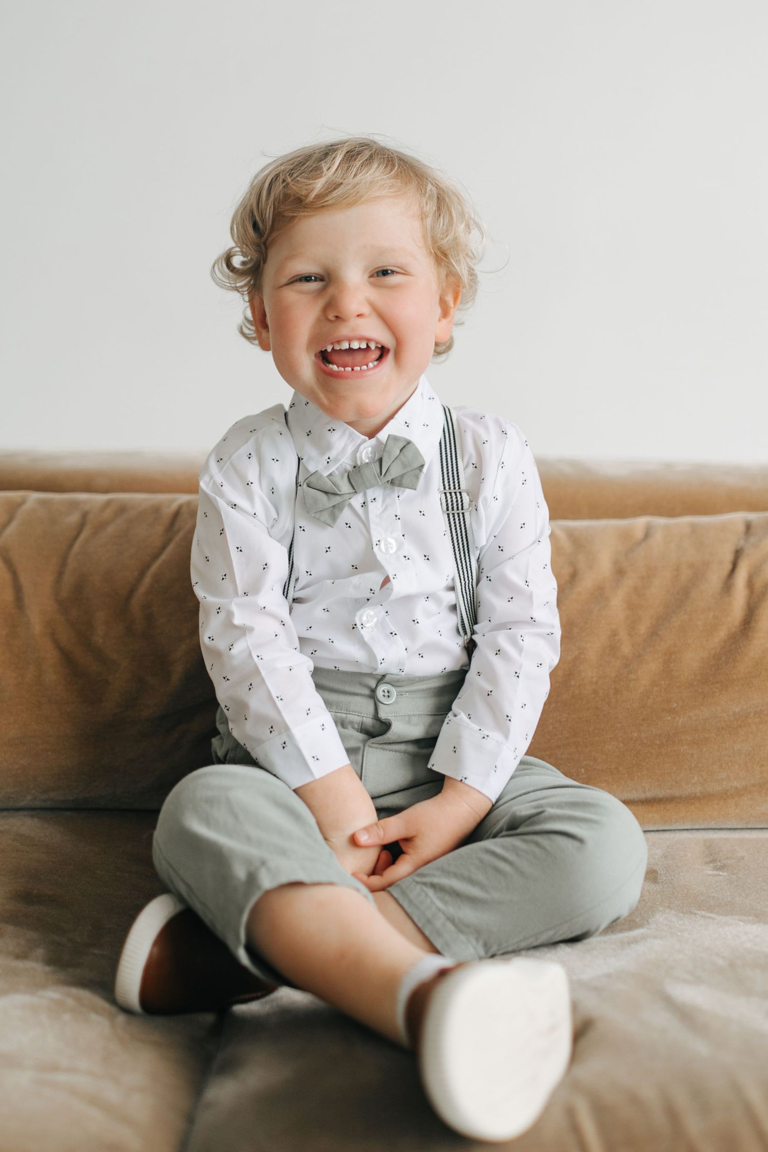 A laughing toddler boy in a patterned white shirt, suspenders, and sage pants sits on a beige couch.