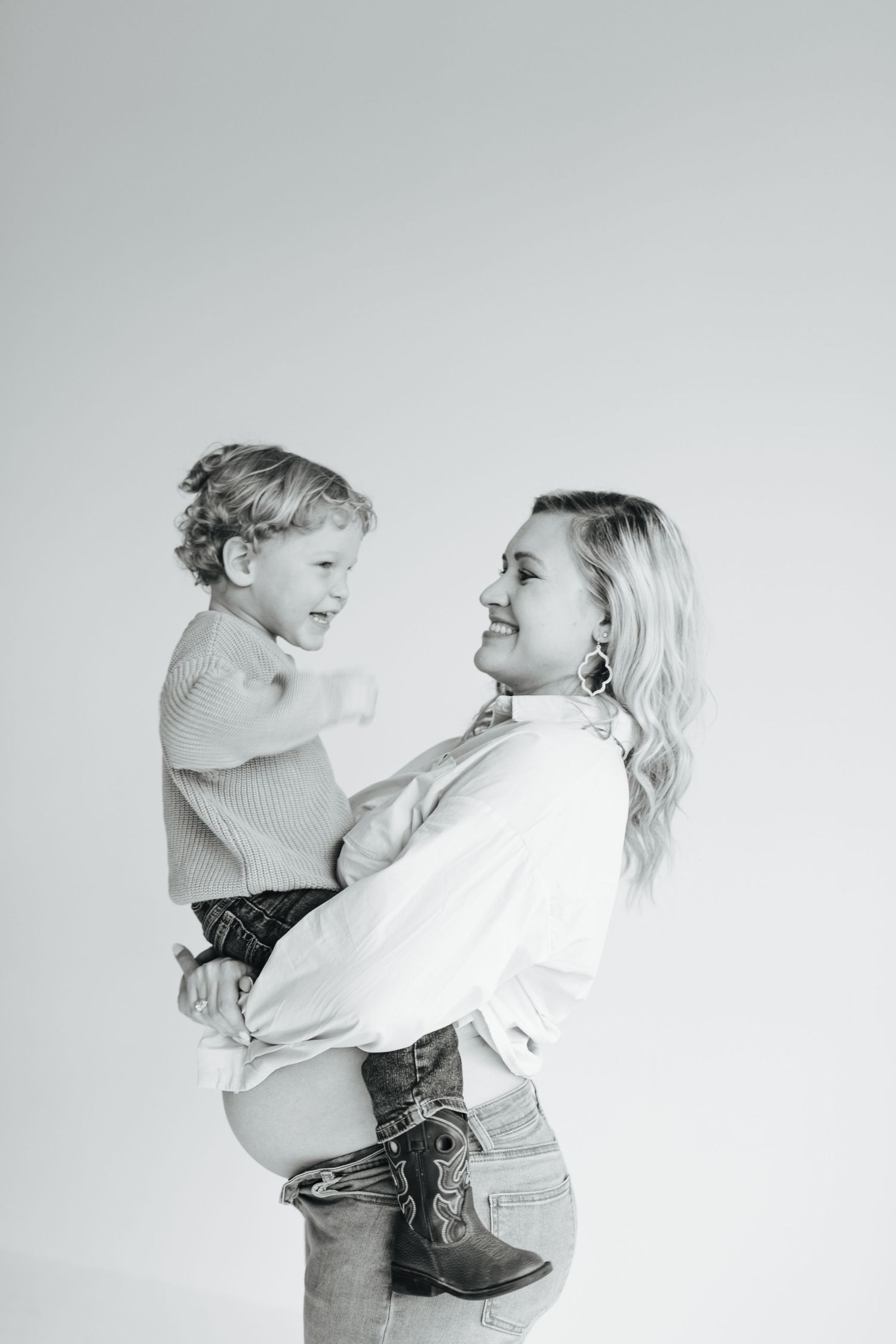 Black and white photo of a pregnant mother holding her laughing toddler son in a studio setting.