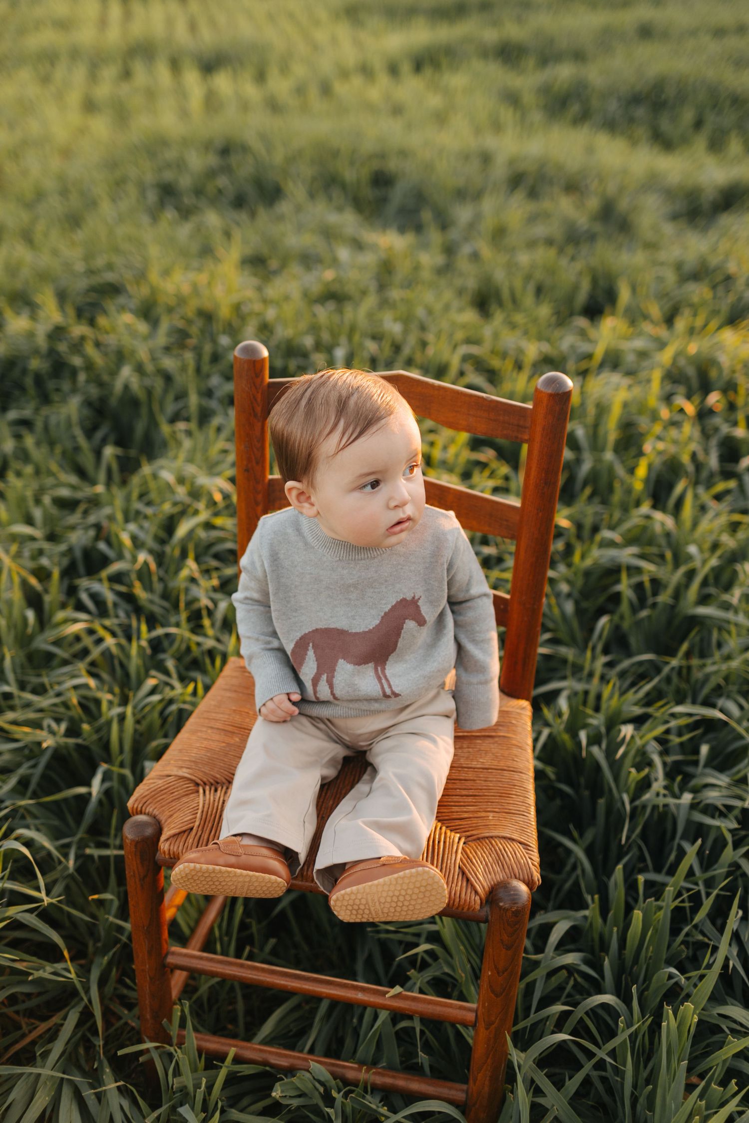 Curious baby in horse-print outfit sits on woven wooden chair surrounded by lush green field at sunset.