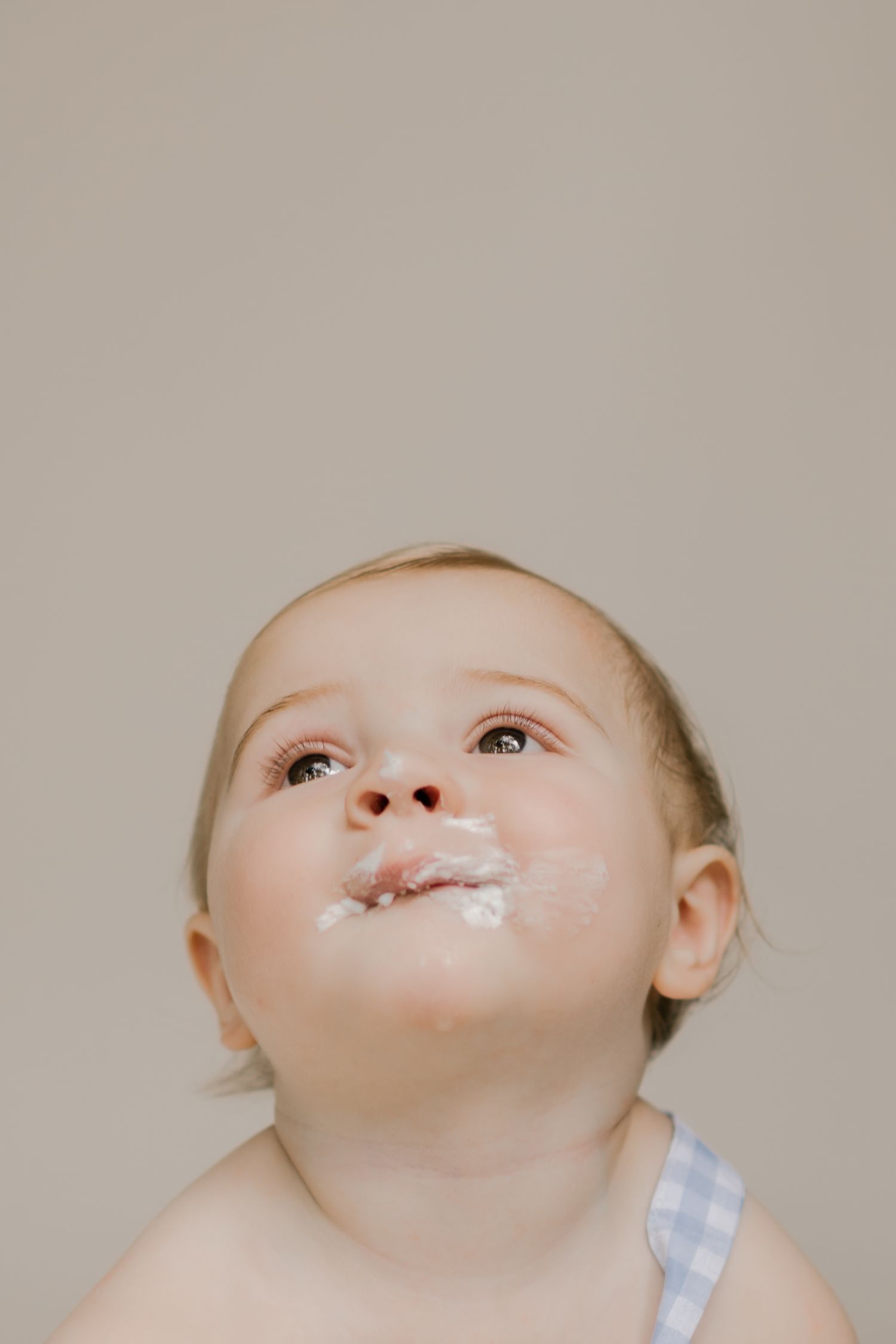 Cute baby with white frosting on face looking up, wearing blue gingham bib against neutral background.