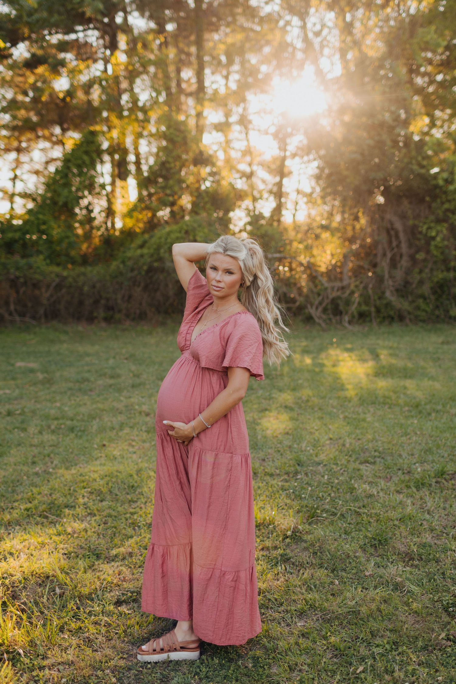 Pregnant woman in pink maxi dress poses in golden sunlight with hand in hair.