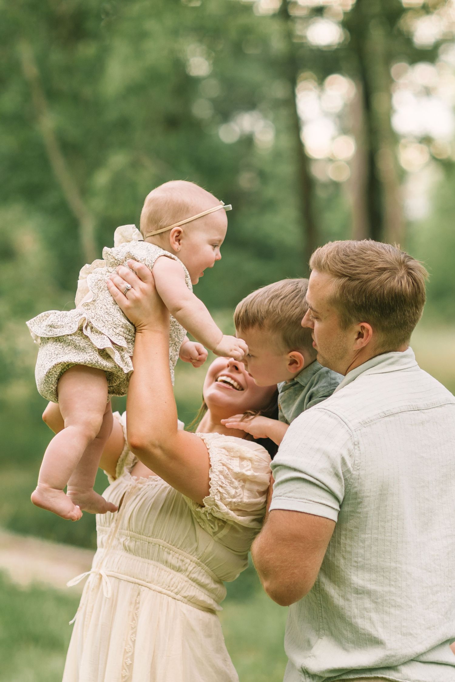 Happy family outdoors, mother lifting smiling baby while father and young son laugh together in a lush green park.