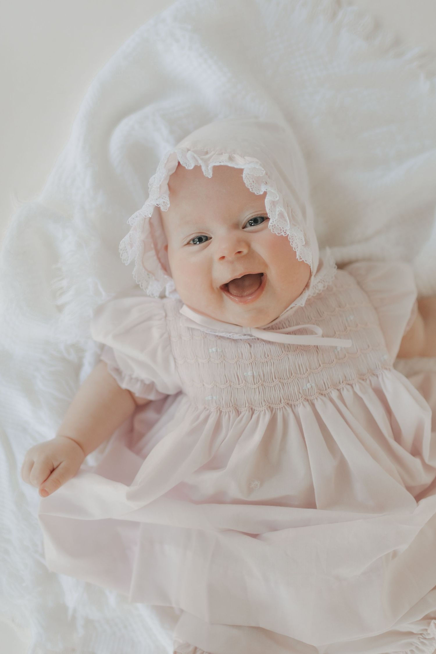 Smiling baby girl in pink smocked dress and lace bonnet lying on white blanket.