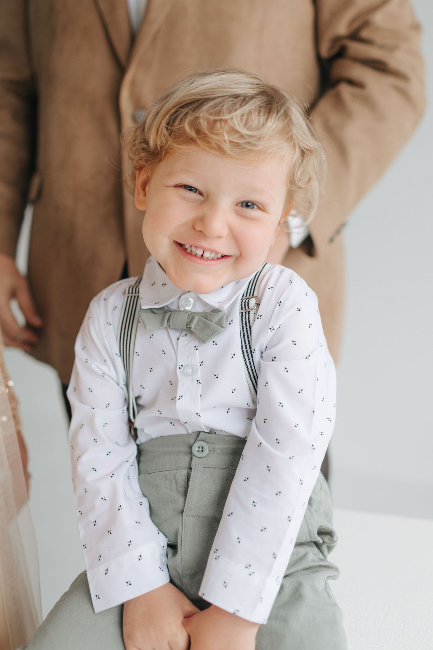 A cheerful blond toddler boy in a white shirt with suspenders and sage green bow tie smiles brightly.