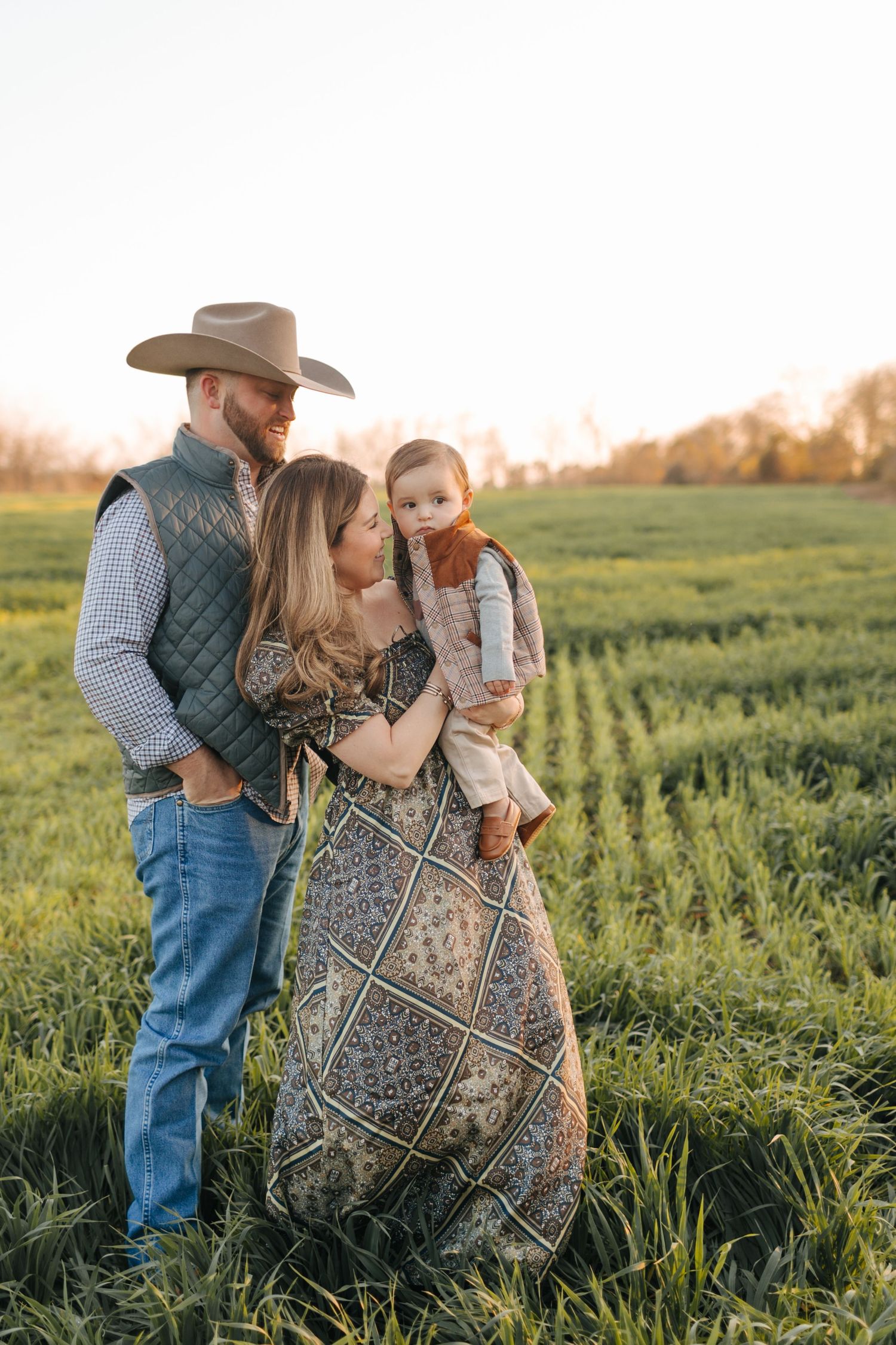 Couple with man in cowboy hat and woman in boho dress holding their baby in a green field at sunset.