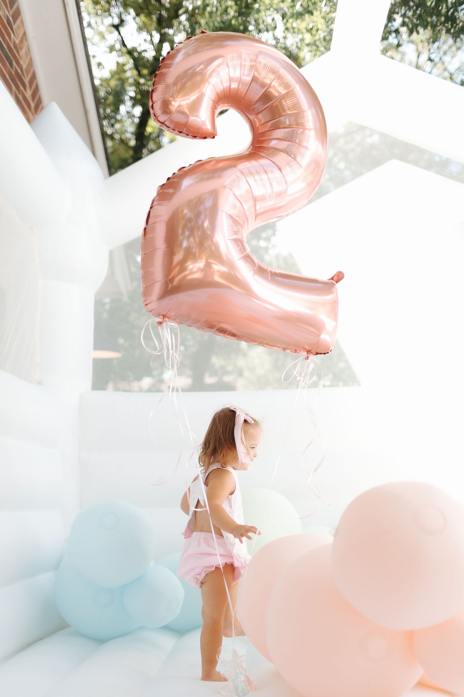 Toddler girl in pink swimsuit plays with pastel balloons in white bounce house at 2nd birthday party.