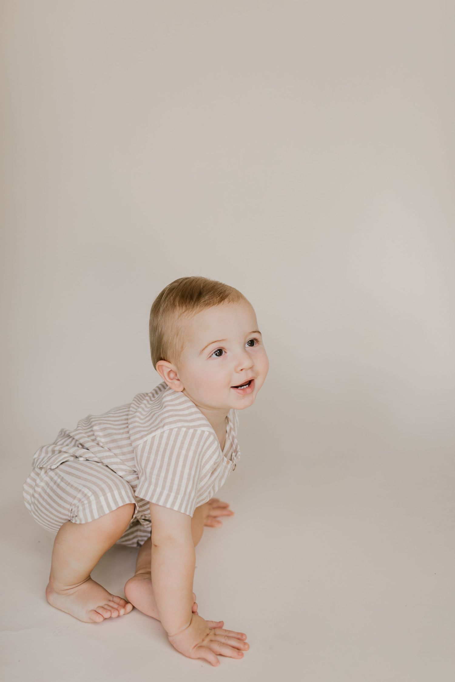 Adorable baby in striped romper crawling on white floor, looking up with a curious smile.