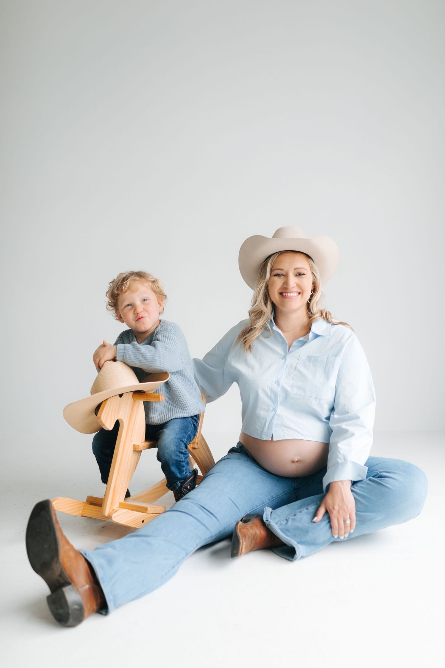 Pregnant woman in cowboy hat sitting on floor with young son on wooden rocking horse in studio.
