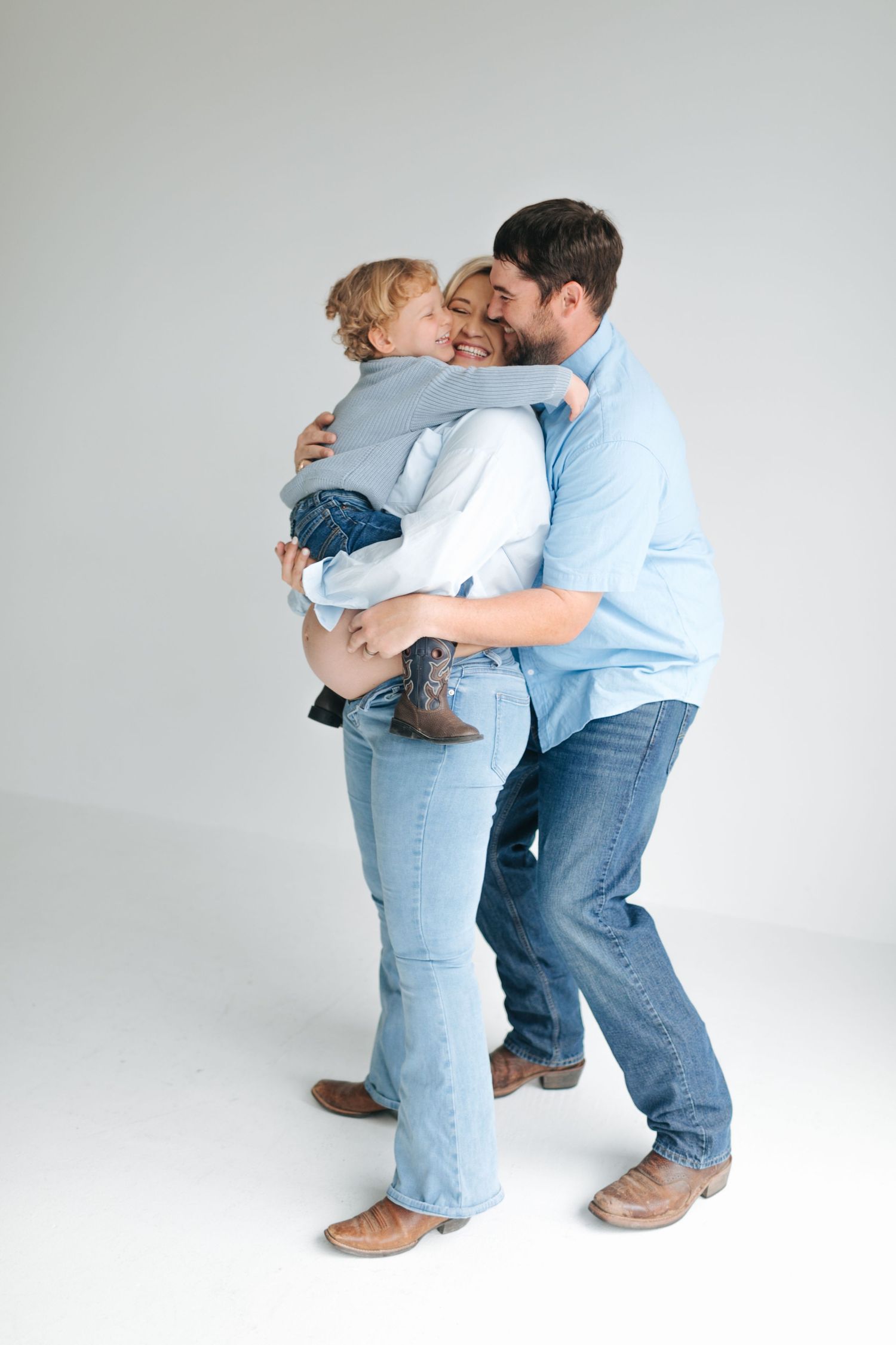 A joyful family in coordinating blue denim embraces together in a bright white photography studio.