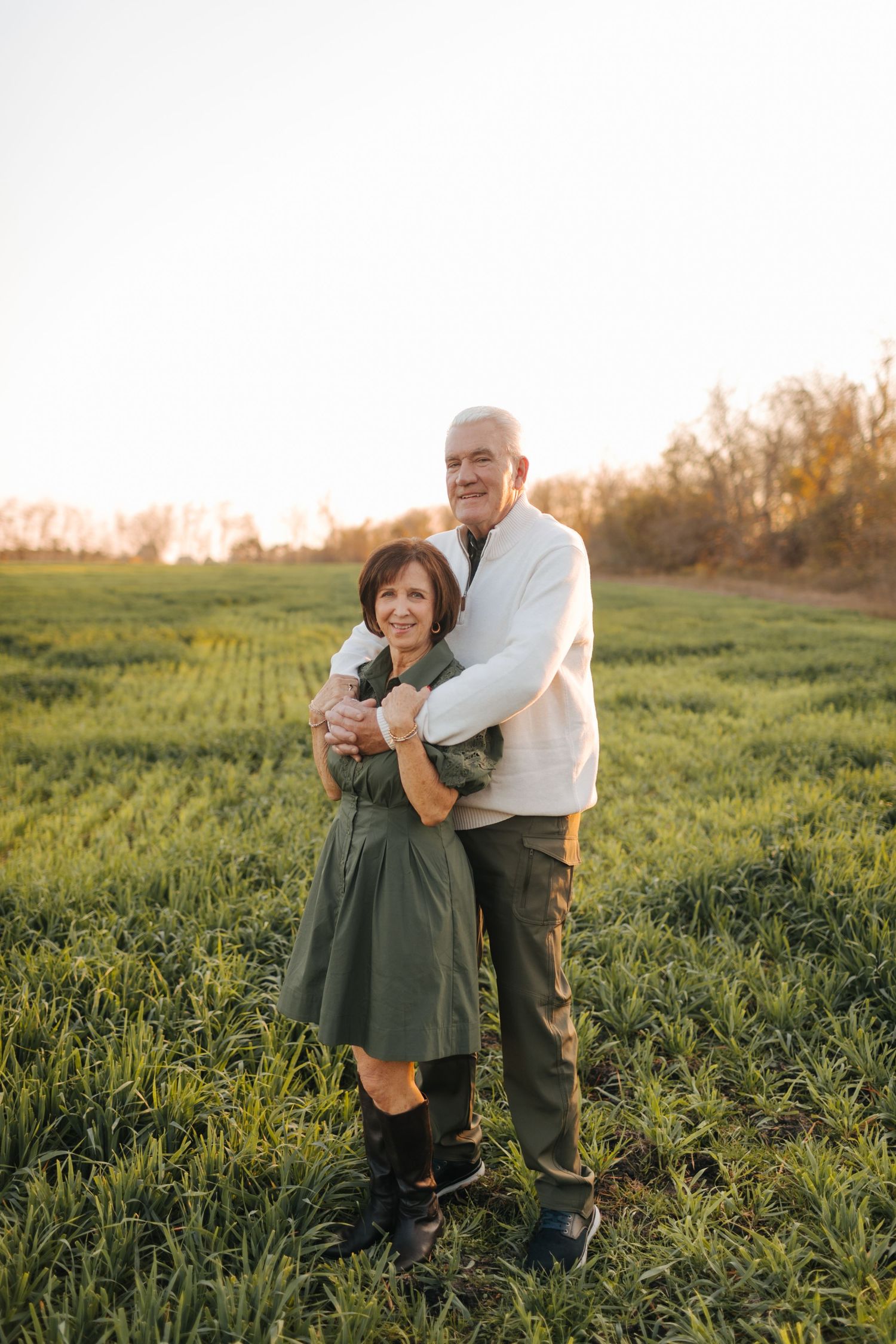 Senior couple embracing in green field at sunset, woman in olive dress with boots, man in white sweater.