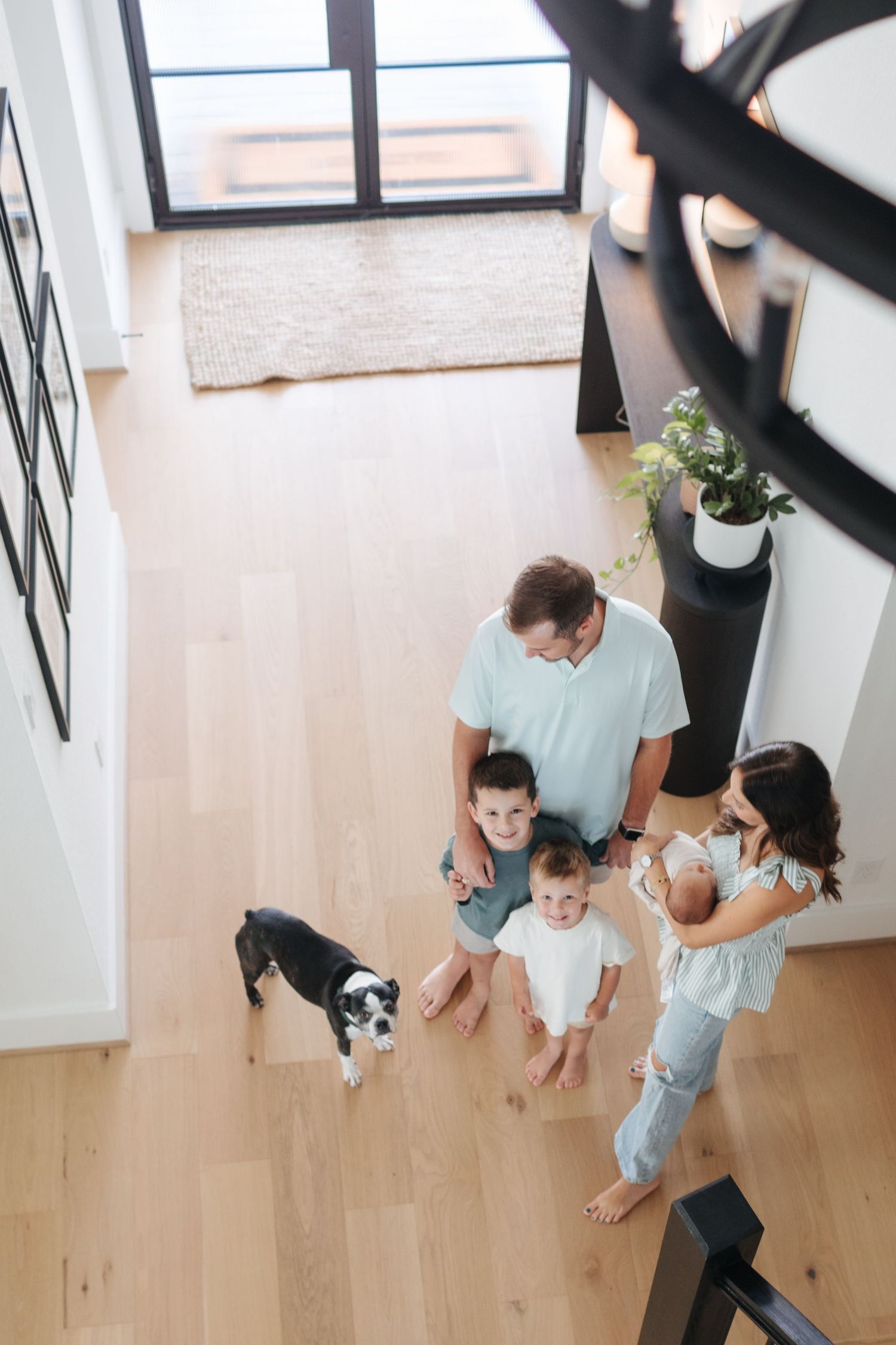 Overhead view of family with dog, toddler, and newborn gathered on light wood floor at home.