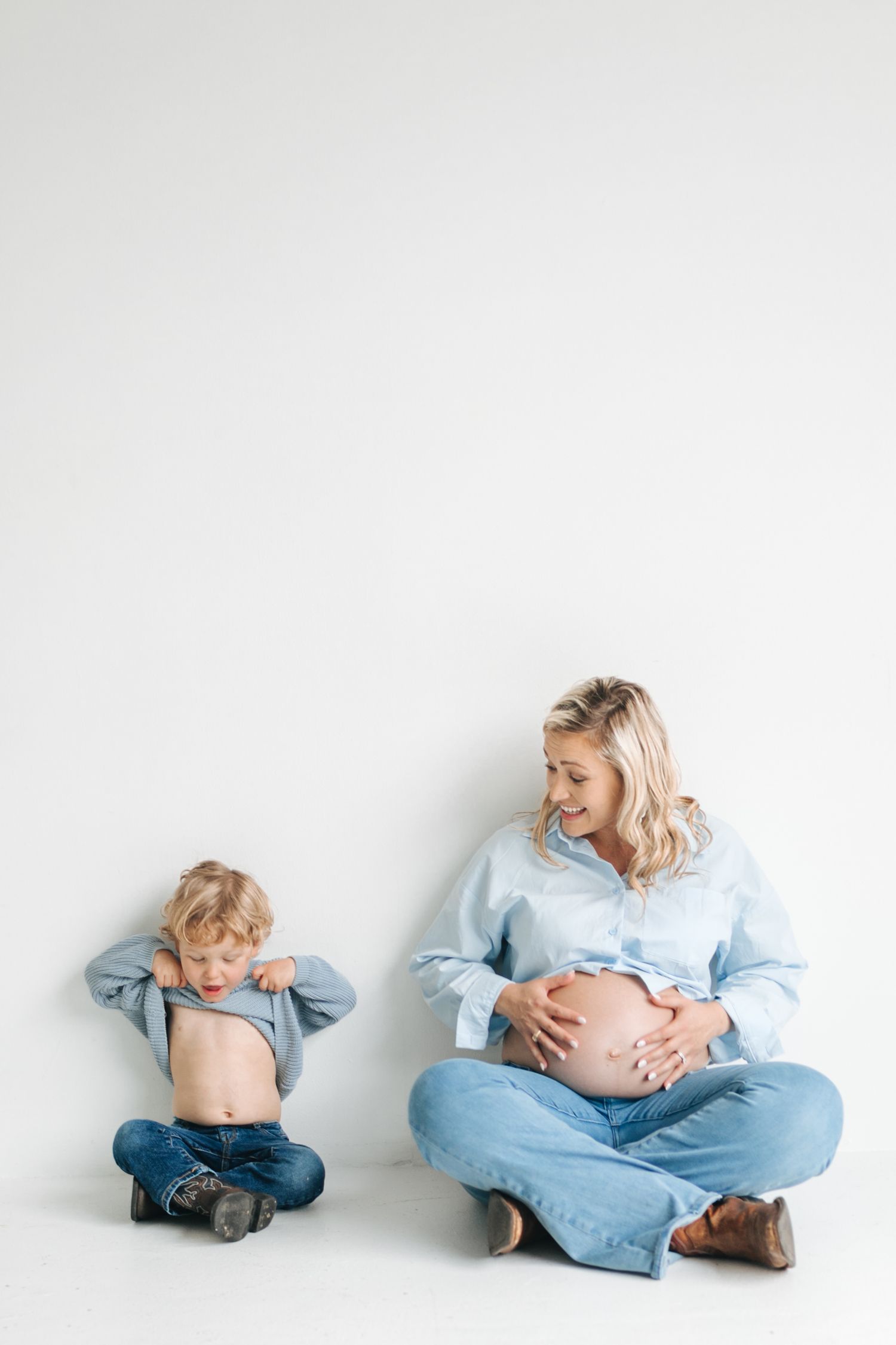 A smiling pregnant woman in blue denim sits beside her toddler son as both show their bare bellies.